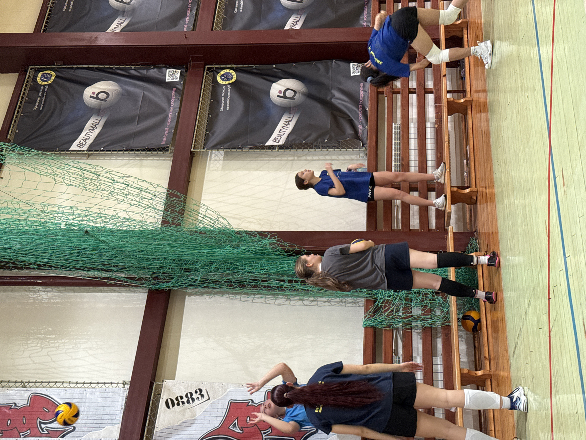 The image depicts an indoor volleyball training session occurring at a sports hall in Sofia, Bulgaria. Six young females are present on a polished green court marked with red and blue lines.
In the upper left, a yellow and blue volleyball is airborne. On the left foreground, a female with long dark red hair in a ponytail, wearing a dark blue t-shirt with "BK 1947", black shorts, white ankle braces, and athletic shoes, stands with her back to the viewer. Behind her, another female in a light blue t-shirt is seen with arms raised, looking upwards, in a setting posture. In the midground, a female with long brown hair in a ponytail, dressed in a gray t-shirt, dark shorts, black leg sleeves, and athletic shoes, holds a yellow and blue volleyball in her left hand, positioned in profile. To her right, another female in a blue t-shirt and black "NIKE" shorts stands on a light wooden bench, observing. In the far right foreground, a female with dark hair in a ponytail, wearing a blue t-shirt, black shorts, white knee pads, and white athletic shoes, is bent over in a low defensive stance, facing away from the camera. A sixth female, partially visible in the upper left background, wears a light blue top.
The background consists of light beige walls with dark brown structural beams and window frames. A green, large-mesh net hangs vertically from the ceiling. Two light-colored wooden benches are situated along the lower wall. Multiple black banners are affixed to the walls, prominently featuring "BEAUTYMALL.bg" as an "ОФИЦИАЛЕН СПОНСОР на волейболен клуб" (Official sponsor of the volleyball club), with a club emblem, QR code, and Bulgarian text "BeautyMall - красотата е споделена" (BeautyMall - beauty is shared). A partially visible banner with "0883" and red graphics is on the left wall. An additional yellow and blue volleyball rests on the floor next to one of the benches.