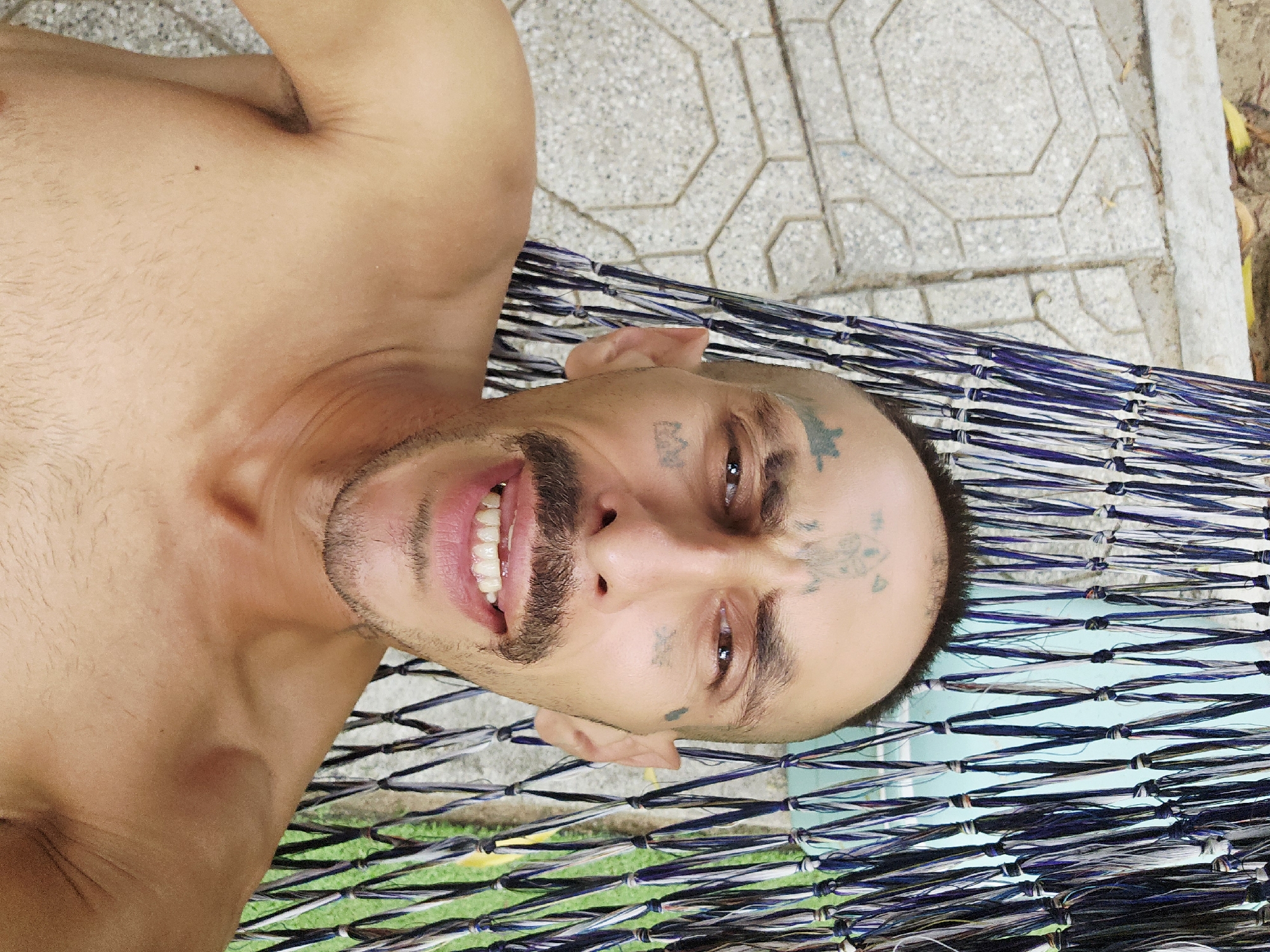 A man lies on a blue and white woven hammock in Cam Lam, Vietnam. The hammock rests on a light grey, hexagonal tiled surface. The man has light skin, short dark hair, and a moustache. Tattoos are visible on his face. He is smiling, exposing his teeth. His right shoulder is visible.