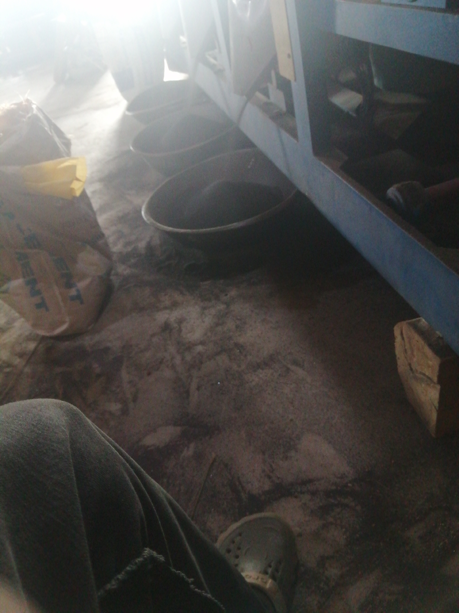The image depicts an indoor scene, possibly a workshop or storage area, with a dusty, gritty floor. In the foreground, a person's legs and feet are visible, wearing dark trousers and a light-colored shoe. To the left, a bag, possibly containing cement, with visible text, lies on the floor. Several dark, circular basins are arranged in a line, filled with a dark, powdery substance. In the background, a blue machine or structure is partially visible, with shelving and various items stored within its compartments. The lighting is bright and diffused, suggesting an overhead light source or natural light entering from a window. The scene appears to be set in a utilitarian environment, potentially related to construction or manufacturing. Without further information or context, it is not possible to definitively determine the location within Jos, Nigeria.