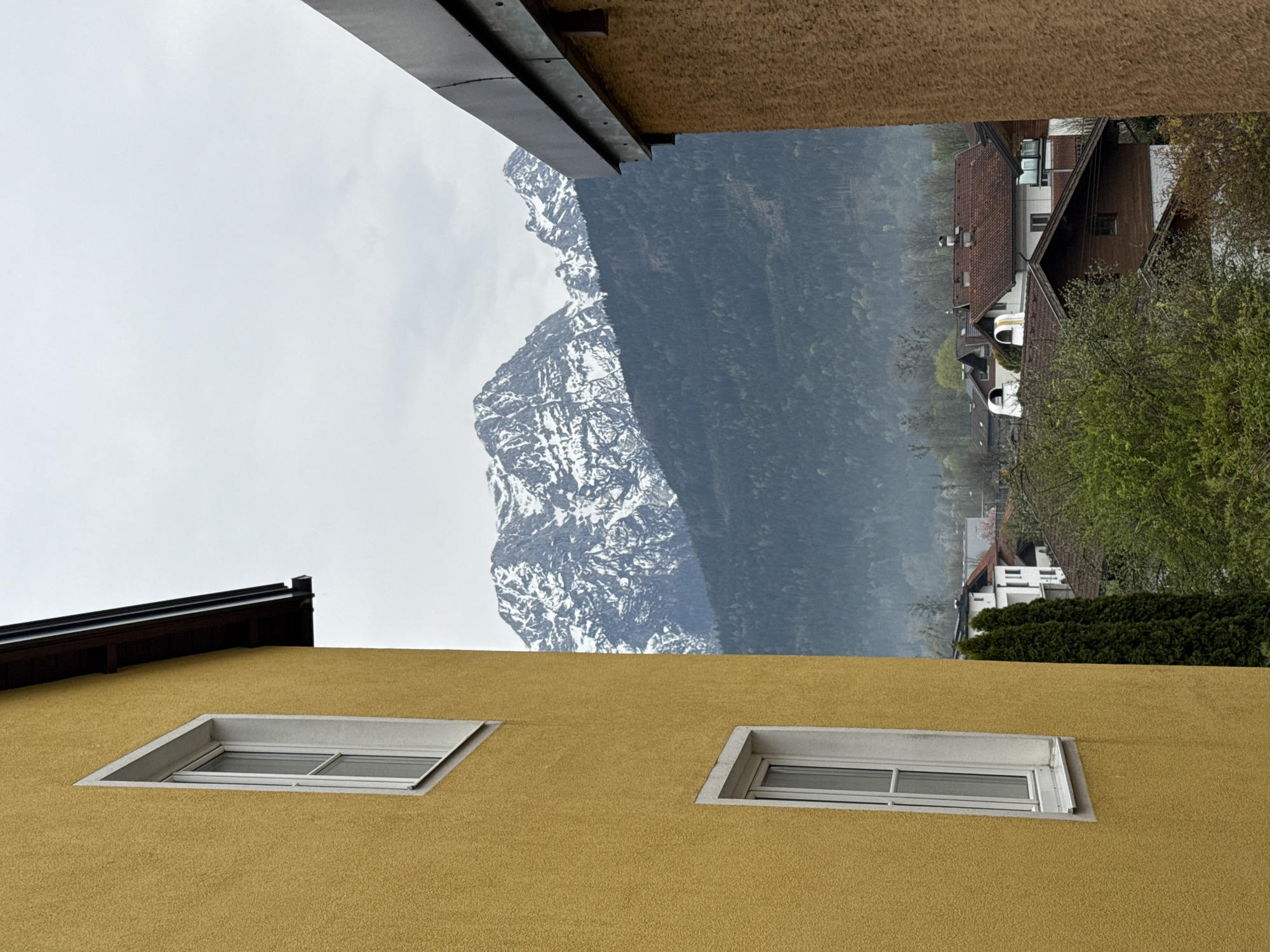 The image shows a view framed between buildings in Moos-Siedlung, Austria, oriented with the ground to the left. A yellow stucco building with two rectangular windows dominates the foreground. Above, a mountainous landscape with snow-capped peaks is visible, shrouded in mist. The background includes houses with red-tiled roofs and patches of green trees. The sky is overcast. No people are visible.