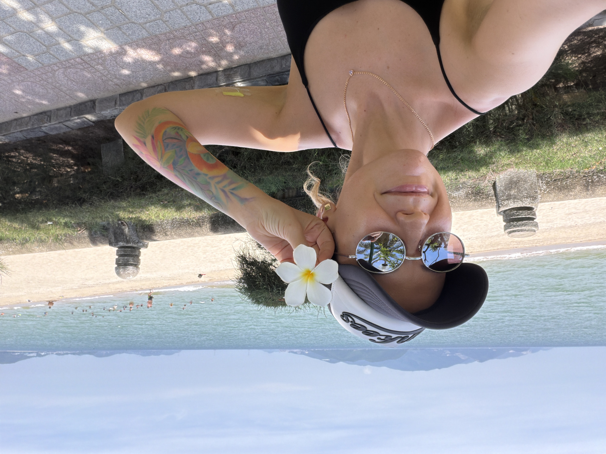 A woman wearing a black tank top, sunglasses, and a black and white baseball cap is holding a white flower with a yellow center to her face. She has blonde curly hair and a colorful tattoo on her right arm. She is standing on a paved walkway next to a line of bushes and stone pillars. In the background, there is a sandy beach with people swimming in the ocean and a distant coastline with mountains. The scene appears to be a beachfront location in Nha Trang Ward, Vietnam.