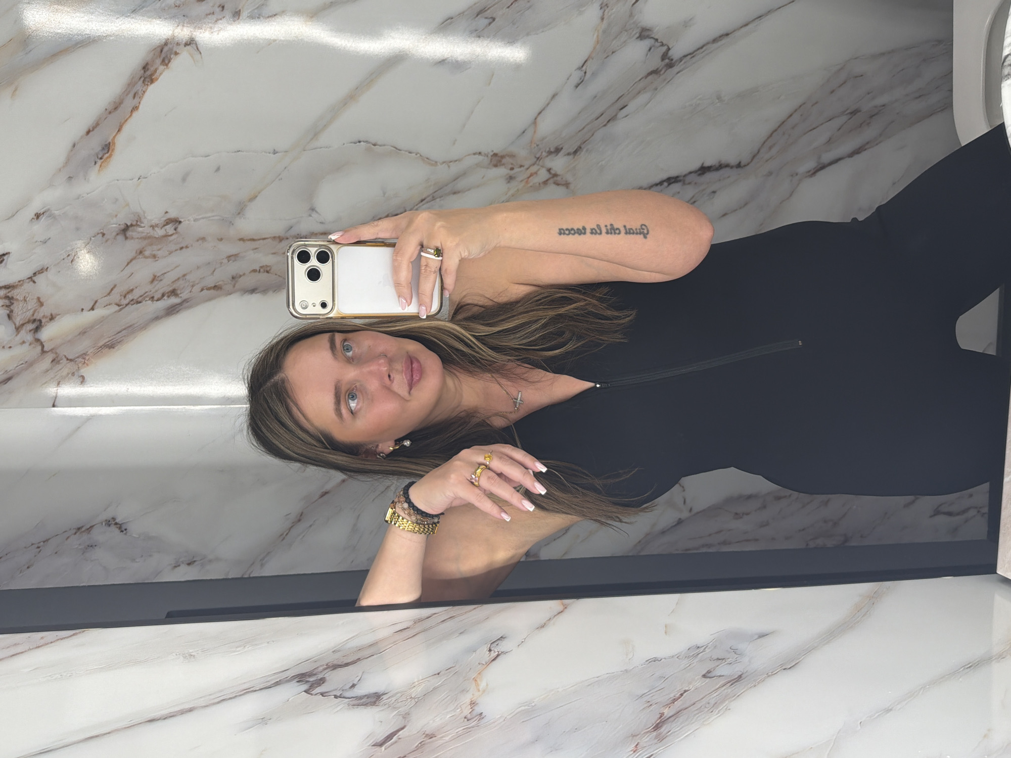 A woman with blond hair is taking a selfie in a bathroom. She is wearing a black jumpsuit and gold jewelry. The bathroom has marble walls and floors. The location is unknown and cannot be determined from the image.