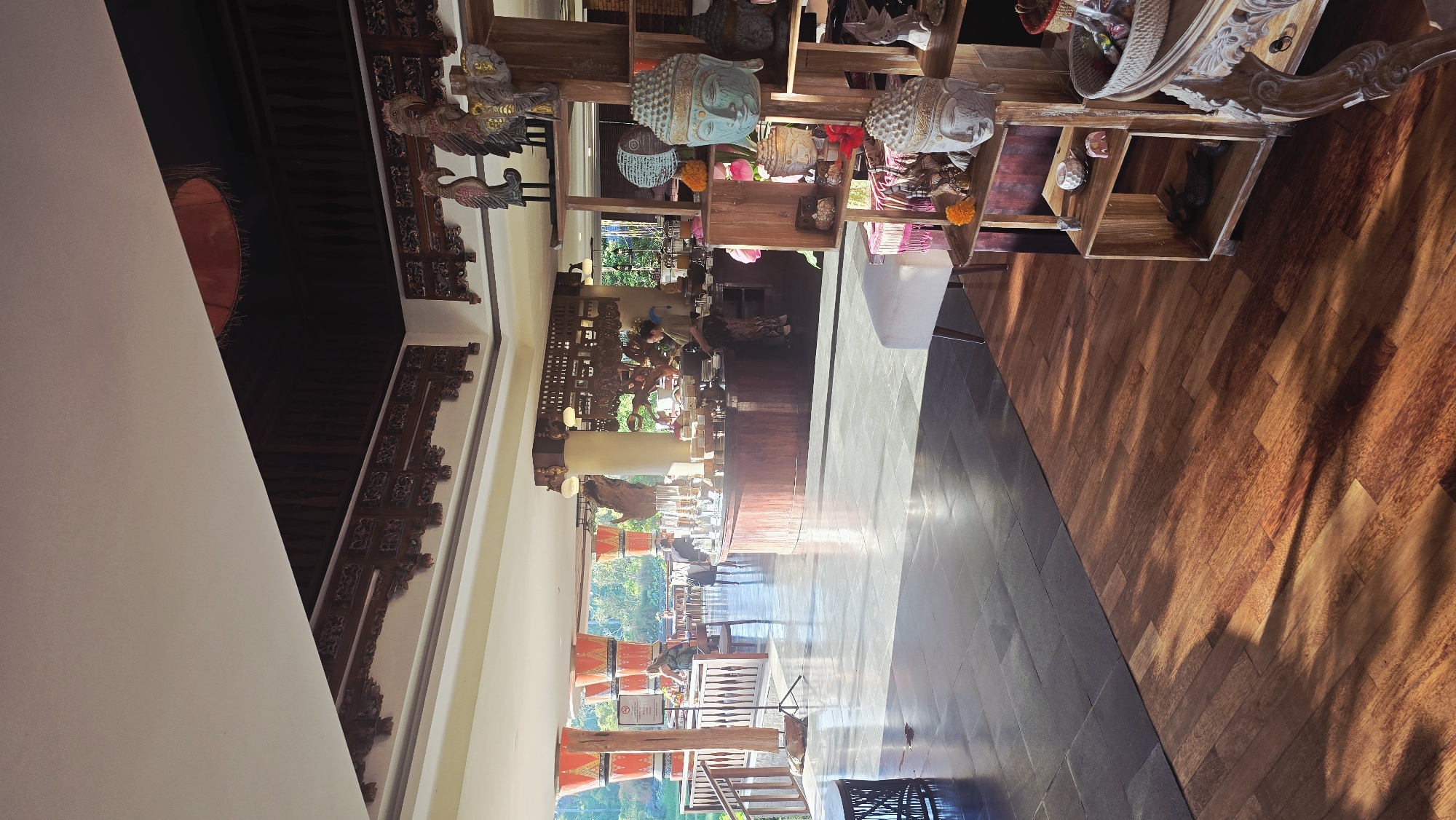 This indoor scene shows a well-lit interior space, seemingly a restaurant or shop, located in Ubud District, Indonesia. In the foreground, there are wooden shelves displaying sculptures, including Buddha heads. The shelves are positioned near an area with a dark, possibly wooden, floor. The background features a long counter with people behind it, likely staff. Beyond the counter, there's an outdoor area with tables and chairs, suggesting a patio or garden setting. The lighting suggests natural sunlight filtering through the outdoor area, creating a bright and inviting atmosphere. Architectural details include ornate woodwork along the walls and ceiling.