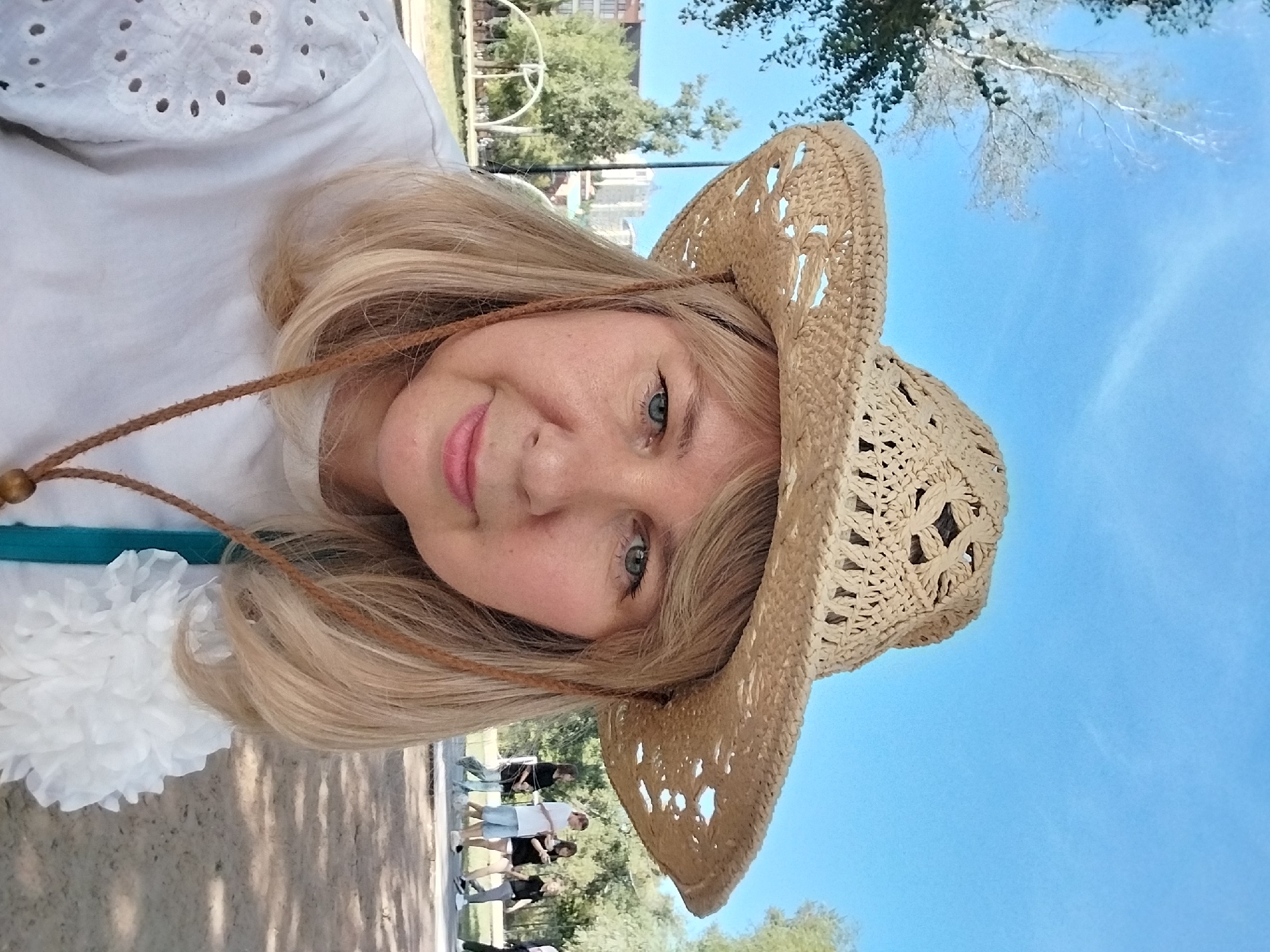 A fair-skinned woman with blonde hair and blue eyes is pictured in a selfie. She is wearing a woven straw cowboy hat and a white shirt with eyelet detailing on the sleeves. A thin brown cord from the hat is visible, ending in a wooden bead. She is also wearing a teal strap, possibly from a bag. The woman has makeup on, including pink lipstick and blush.
The background suggests an outdoor urban park setting. Trees with green foliage are present, and in the distance, a multi-story building with light-colored walls is visible. Several people are walking in the background, appearing to be on a paved path. The ground in the foreground is sandy and uneven, with shadows cast on it. The sky is bright blue with a few wispy white clouds.
The image appears to have been taken on a sunny day. The location is identified as the Orenburg region of Russia, specifically near a children's playground and sports ground. The time appears to be daytime, likely late morning or afternoon, given the lighting conditions. There is no explicit action or interaction occurring between subjects within the frame, other than the selfie being taken by the woman.