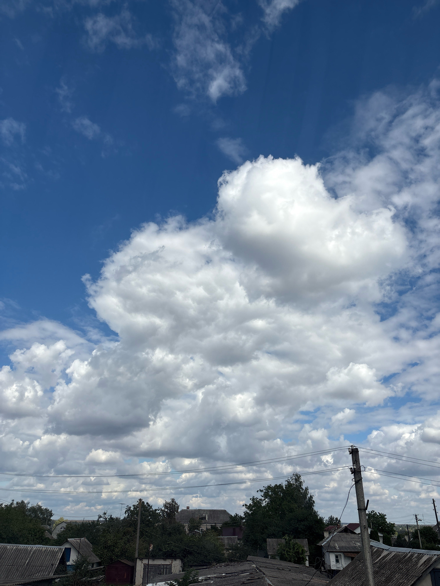 A vertical, eye-level shot depicts a partly cloudy sky over a village. The sky is a deep blue with scattered white cumulus clouds, some of which are large and fluffy, while others are wispy. Sunlight streams through the clouds, creating visible rays. Below the sky, the upper parts of houses, trees, and utility poles are visible. The houses have visible roofs, some with visible chimneys. Trees are dense and green, obscuring much of the lower parts of buildings. Utility poles and wires are present in the foreground and mid-ground. The overall impression is of a rural or semi-rural landscape under a dynamic sky. This scene is in Lanivtsi, Ukraine.