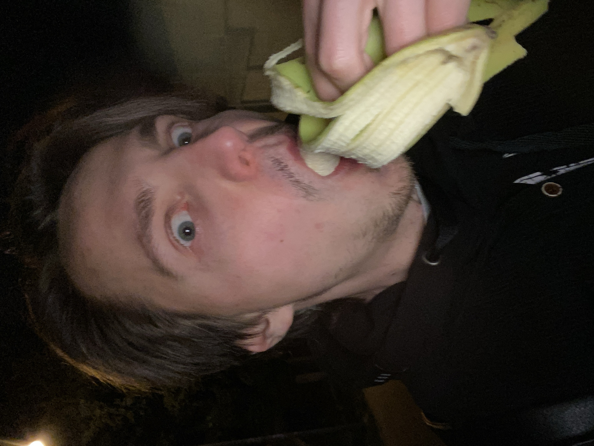A man with light skin, light brown hair, and stubble is eating a banana peel. The man is wearing a black hoodie. The location is not evident from the image.