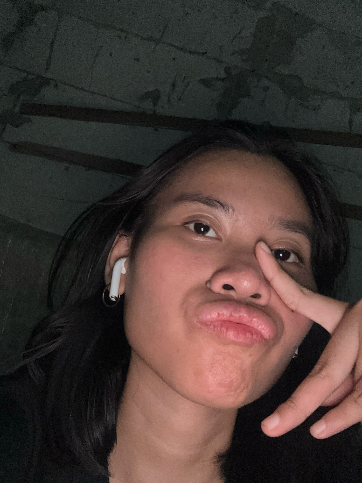 A young woman with dark hair is taking a selfie. She is wearing white earbuds and a hoop earring. She has her right index finger to her nose and is puckering her lips. The background appears to be dark and possibly a ceiling or textured wall. The image is taken at night in a dimly lit indoor environment. The user is in East Jakarta, Indonesia.