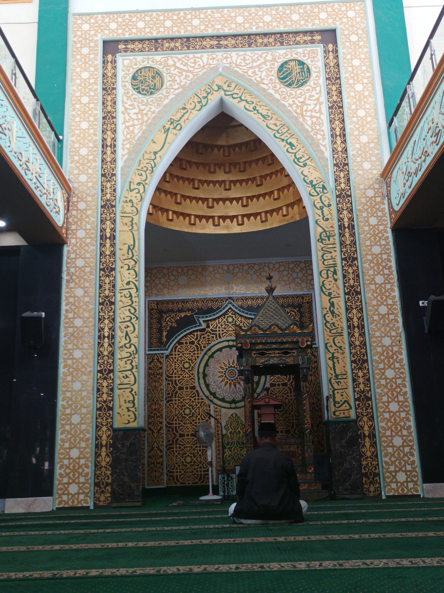In the city of Nganjuk, Indonesia, a man is seen from behind, sitting on the green carpeted floor of a mosque, facing a Mihrab. The Mihrab is elaborately decorated with intricate patterns in gold and teal. The man is wearing a dark outer garment and white trousers. He appears to be engaged in prayer or meditation. The mosque's interior features tall, ornate columns and geometric motifs on the walls. A fan stands to the left of the Mihrab, and what appears to be a small pulpit or decorative structure is positioned within the Mihrab. The overall ambiance is one of reverence and architectural splendor.