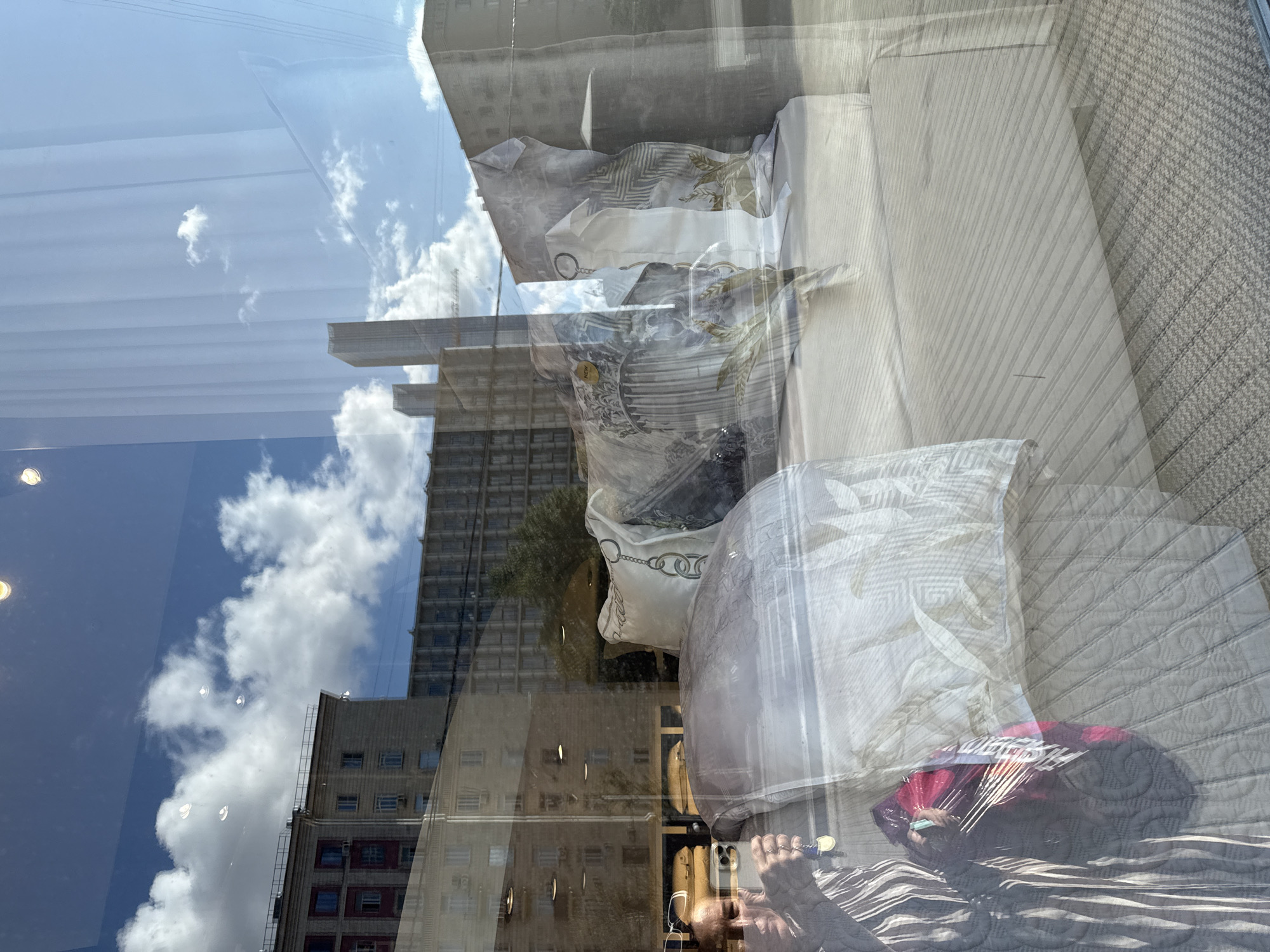 This image captures a reflection in a storefront window, showing a display of bedding and pillows. A person is visible on the left, holding a smartphone and keys, appearing to be taking a selfie. The display includes a neatly made bed with several pillows, one of which has a detailed pattern featuring chains and architectural elements. The bedding itself has a light-colored, patterned fabric.
Through the reflection, we see a city street scene with buildings and a cloudy blue sky. The buildings appear to be apartment blocks, typical of Moscow's Soviet-era architecture. Specifically, the building on the left, with its red-brick lower floors and cream-colored upper floors, resembles buildings found in areas like Arbat or other central Moscow districts. The presence of multiple stories, balconies, and uniform window placements are characteristic of residential buildings in Moscow. The sky is bright with fluffy white clouds, suggesting a clear, sunny day. The overall impression is a snapshot of everyday urban life in Moscow, with the storefront display offering a contrast to the exterior environment.