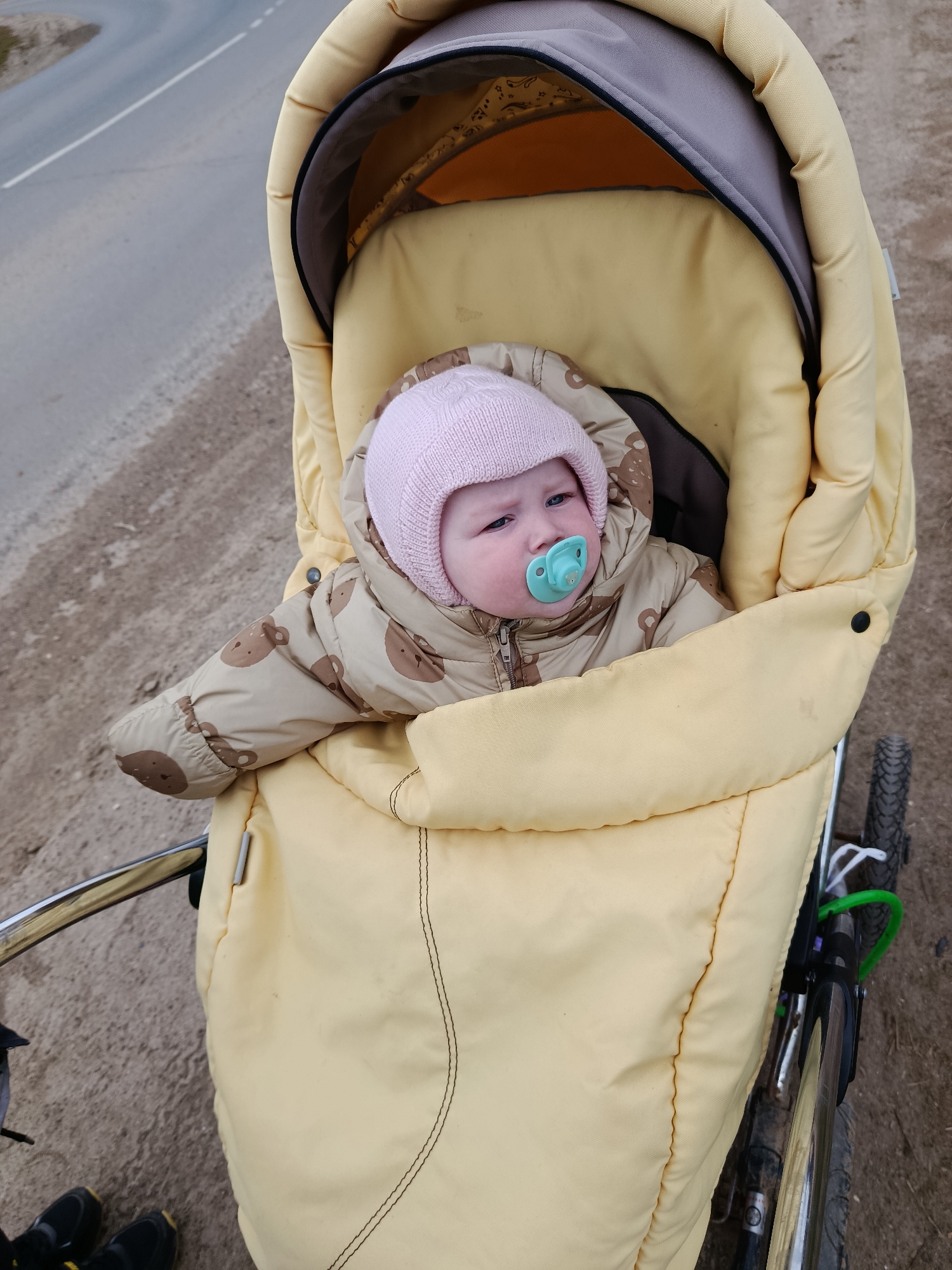 A baby is depicted in a close-up, slightly high-angle shot, seated in a yellow pram-style stroller outdoors. The infant wears a light pink knitted hat and a hooded, padded jumpsuit in a light brown or tan color, featuring a pattern of small brown bear faces. A bright turquoise pacifier is in the baby's mouth, and its eyes are open, looking slightly to the right with a somewhat neutral or pensive expression.
The stroller's interior is well-padded and yellow/light beige, with a grey fabric lining visible under the canopy. Parts of the stroller's metal frame, which appears to be chrome, are visible on the left and bottom, along with a black wheel on the right side of the frame.
In the background, a grey asphalt road with a white dashed line is visible to the left, suggesting a travel lane. A dirt or gravel shoulder runs alongside the road and underneath the stroller. The lighting is diffused, indicating daytime, possibly an overcast sky, with no harsh shadows. The baby's warm attire suggests cool weather. The scene captures a quiet moment during an outdoor outing in Фоки, Russia. No discernible text is visible within the image.