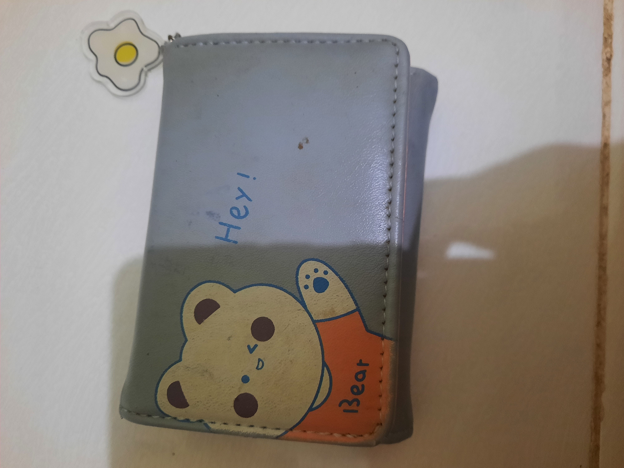 A close-up, high-angle shot shows a light gray tri-fold wallet with a cartoon bear illustration on the left half. The bear is light yellow with brown ears and eyes, wearing an orange shirt. The word "Bear" is written in blue beneath the bear. Blue lettering on the right half of the wallet reads "Hey!". A small, white, cartoon fried egg sticker with a yellow yolk is attached to the upper right corner of the wallet. The wallet rests on a white surface, with a subtle shadow cast to the right. The image is taken at the Lhokseumawe City Square, Lhokseumawe, Indonesia.