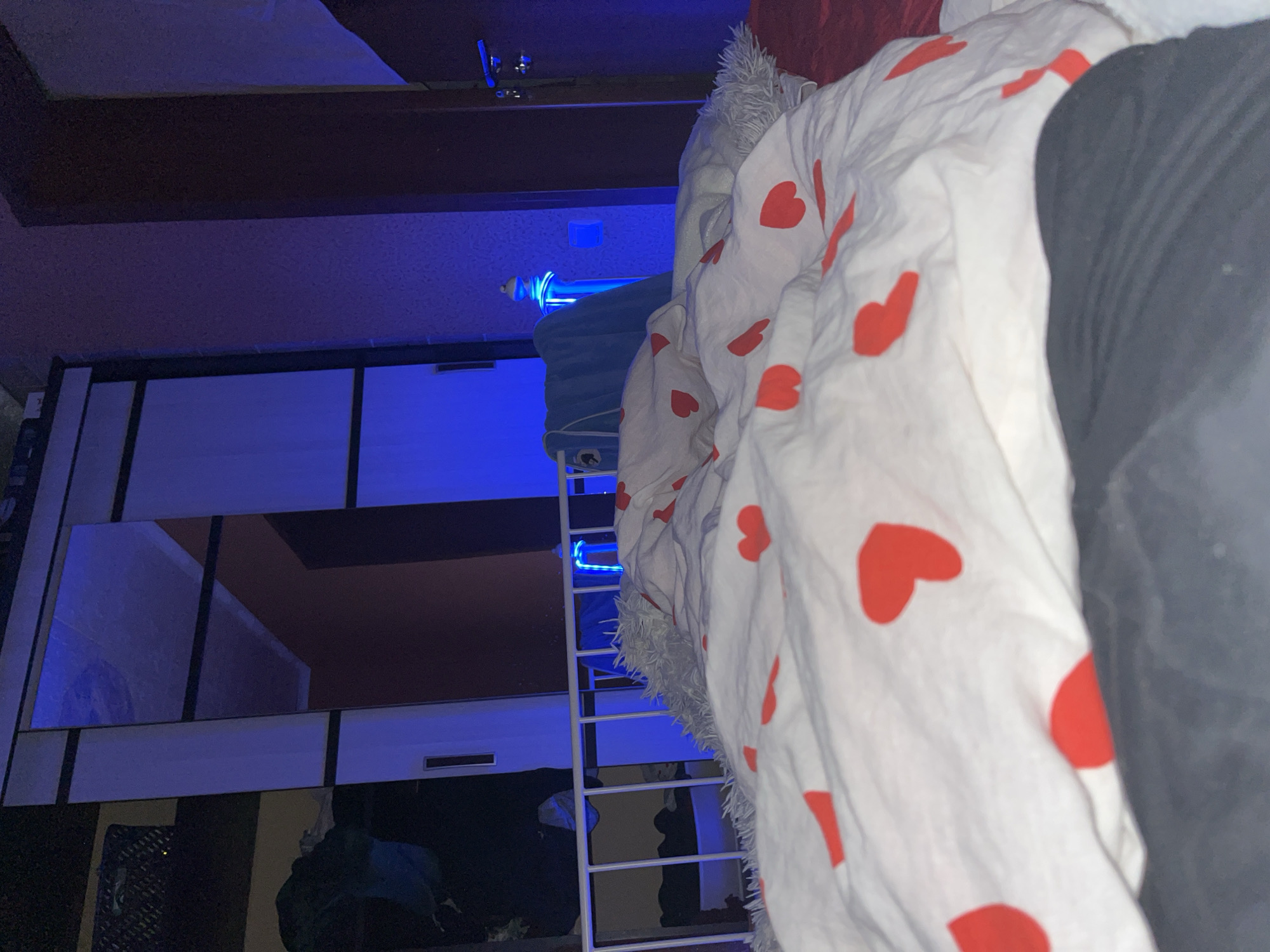 The image depicts an interior scene, likely a bedroom, with a focus on a rumpled bed covered in white bedding with red heart patterns. The lighting is predominantly blue, creating a moodistic atmosphere, with some areas illuminated by a more natural or yellow light.
In the background, a large closet with mirrored doors is visible, reflecting the blue light and parts of the room. To the right of the closet, a dark wooden door with a handle is present. A white railing, possibly part of a bed frame or a decorative element, separates the bed from the background.
Several objects are scattered around the room, including a light blue towel draped over the railing and a fuzzy grey blanket or throw near the head of the bed. The overall impression is one of a lived-in space, possibly a young person's room, given the heart-patterned bedding and the atmospheric lighting.
The specific location in Shakhty, Russia is not determinable from the image itself. However, the scene could be consistent with residential interiors found in urban areas of Russia.