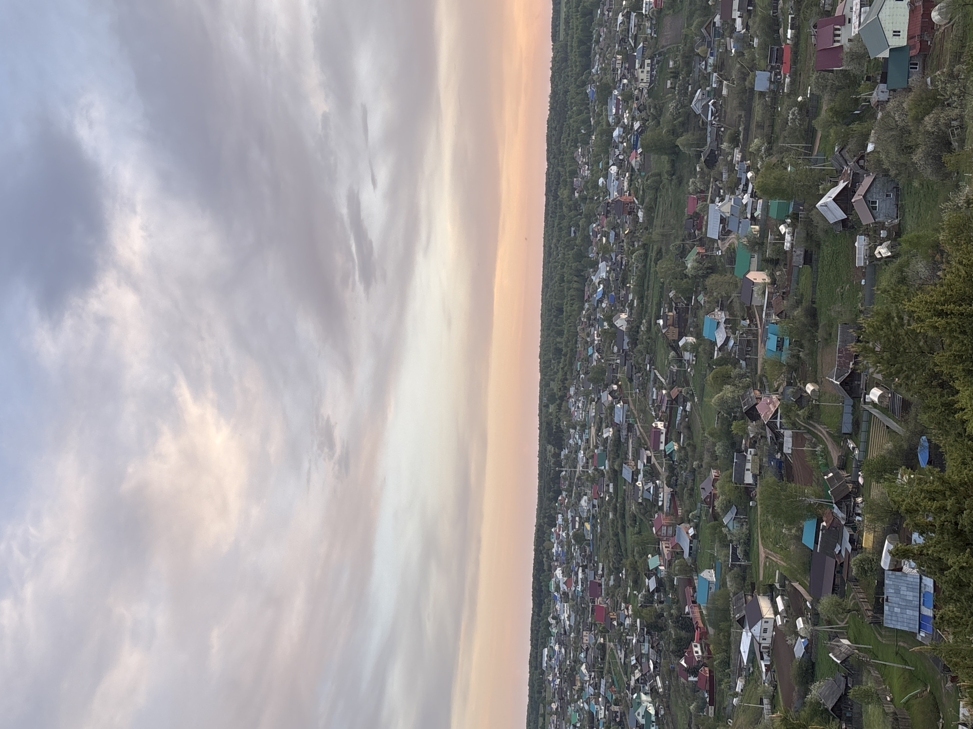 The image depicts an aerial view of a residential area in Прикамский, Russia, at dusk or dawn. The sky, on the left side of the frame, is filled with clouds tinged with soft pastel hues of pink, purple, and grey. A thin, illuminated band of warm light stretches vertically between the sky and the residential area, suggesting the position of the rising or setting sun.
The residential area on the right side of the frame is densely packed with individual houses and structures, interspersed with green trees and vegetation. The buildings exhibit a variety of roof colors, including red, blue, green, and grey. Narrow roads and pathways wind through the settlement. The scene appears calm and still, with no visible human activity.