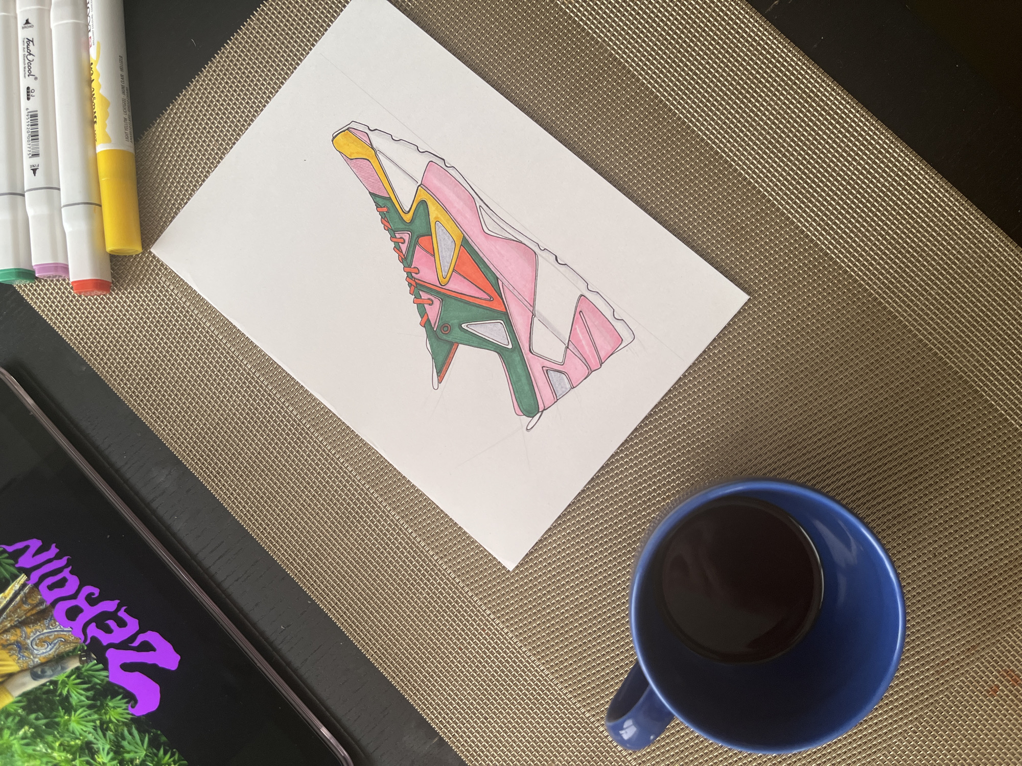 An overhead shot captures a creative workspace featuring a partially colored sneaker drawing, art supplies, a tablet, and a beverage. The scene is set on a textured, golden-brown mat, with a dark surface visible underneath on the left.
The central focus is a white sheet of paper displaying a detailed drawing of a sneaker. The design is abstract and geometric, partially colored in vibrant shades of pink, yellow, dark green, orange, and light grey, outlined in black. Pencil lines are visible, indicating the drawing is in progress.
To the left of the paper, three "Touch Cool" alcohol markers are neatly arranged, their caps revealing green, orange, and yellow colors. Text on the markers includes "Touch Cool," "Broad," "Fine," and "LIGHT YELLOW / JAUNE CLAIR / HELLGELB" on the yellow marker. Numerical codes like "0 21" and a barcode are also visible.
In the bottom left, part of a tablet or smartphone screen is visible, displaying a vibrant image. The screen features large, stylized purple text that appears to read "ZEROPIN" (possibly reversed or highly stylized), set against a backdrop of green leafy material (possibly cannabis) and a yellow/gold patterned fabric.
A dark blue ceramic mug filled with a dark liquid, likely coffee or tea, is positioned in the bottom right corner of the frame. The overall impression is one of a calm and focused creative session, possibly at a desk or table, with elements of art, digital media, and a beverage.