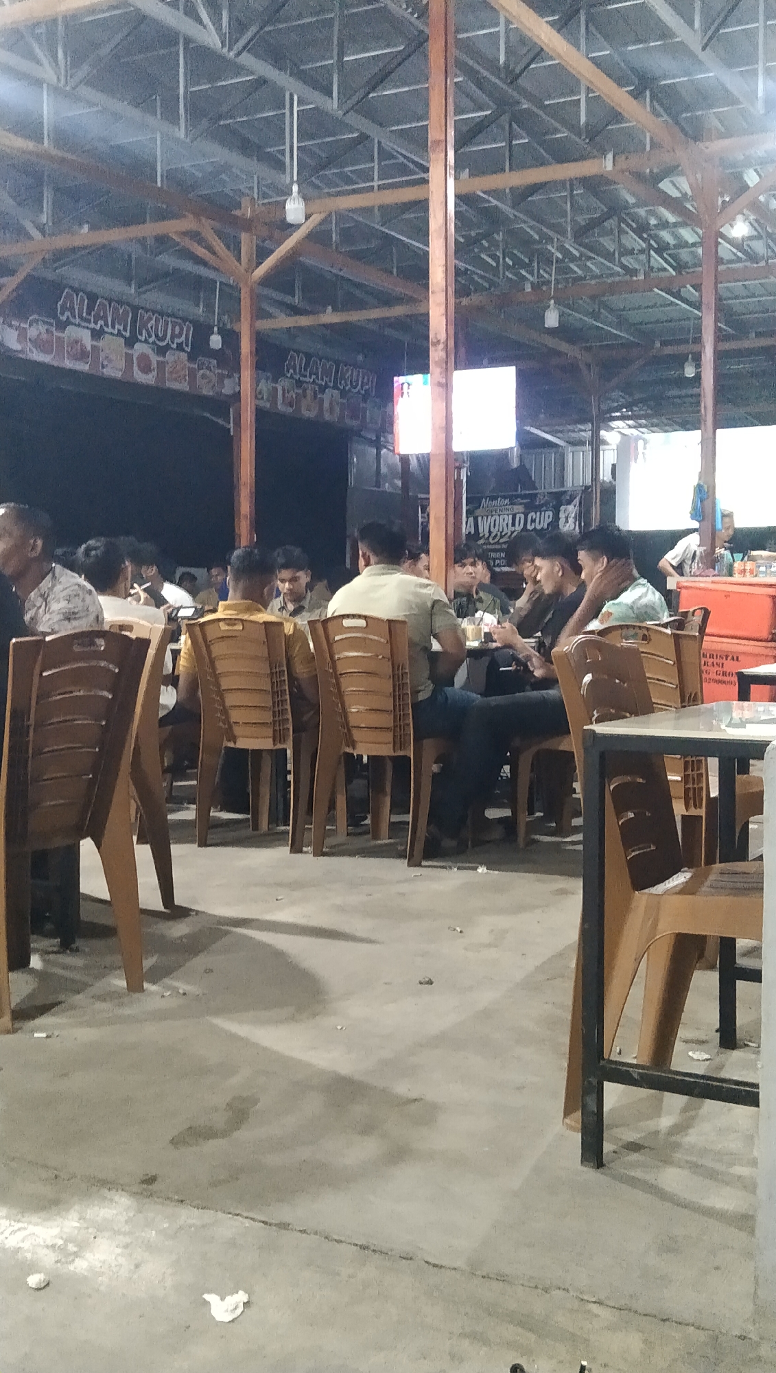 This image depicts a lively outdoor seating area, likely a cafe or restaurant, with people gathered at tables. The setting appears to be a covered space with a metal roof supported by wooden beams, suggesting an open-air or semi-outdoor environment. Several tables with plastic chairs are arranged on a concrete floor.
In the foreground, several empty brown plastic chairs are visible, some casting shadows on the ground. In the background, groups of people are seated at tables, engaged in conversation and dining. Some individuals are facing away from the camera, while others are seen in profile or from the side.
A prominent banner with the text "ALAM KUPI" is displayed, indicating the name of the establishment. Another banner visible in the background advertises "A WORLD CUP," suggesting a sports viewing event. There is a large screen displaying an image, likely part of the World Cup broadcast.
The lighting suggests it is evening or night. The atmosphere seems relaxed and social, with people enjoying food and company. The presence of drinks on the tables and the general arrangement of the seating suggest a casual dining or gathering spot. Visible text on other signs includes "Monton," "Nonton," and "KRISTAL KANI G-GRO." The overall impression is a popular local spot, possibly a warung kopi or simple restaurant, buzzing with activity.