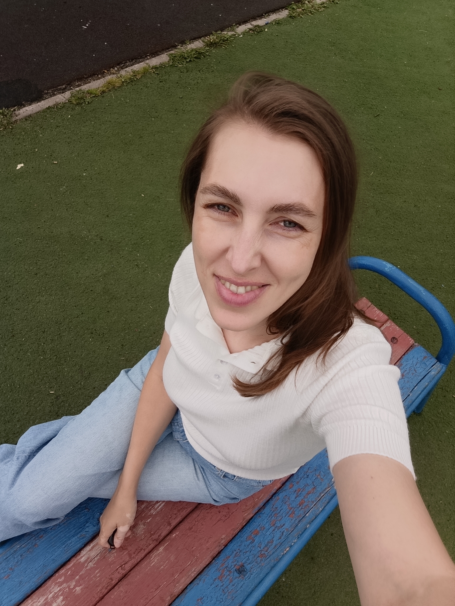 A woman with medium-length brown hair, wearing a white ribbed polo shirt and light blue jeans, is sitting on a colorful wooden bench outdoors. The bench has red and blue painted slats and blue metal supports. She is smiling at the camera, her eyes are blue, and she has light-colored skin with some freckles. Her left hand is resting on the bench, holding a small black object. The ground around the bench is covered in artificial turf, colored green. To the left of the turf, there is a dark asphalt surface with a thin strip of grass separating it from the turf. The scene appears to be in a public outdoor area, possibly a park or playground, located in Chaikovsky, Russia.