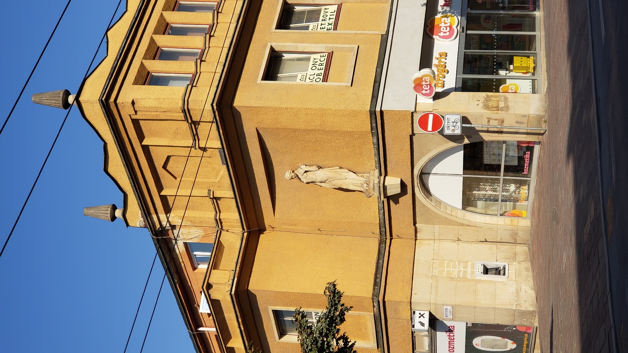 The image shows the facade of a yellow building in the city of Prešov, Slovakia. The building has a decorative cornice and architectural details. A statue of a woman in a flowing robe is mounted on the facade. Above the statue, there is an arched window with display items visible inside. To the right of the arched window, there are shopfronts with signage indicating a "Teta drogéria" (Teta drugstore). There is a red and white circular sign indicating "No entry" and a smaller sign below it with a bicycle symbol and the word "okrem" (except). In the foreground, a paved sidewalk and a street are visible. The sky is clear and blue. The overall scene depicts a street view of commercial establishments in a European city.