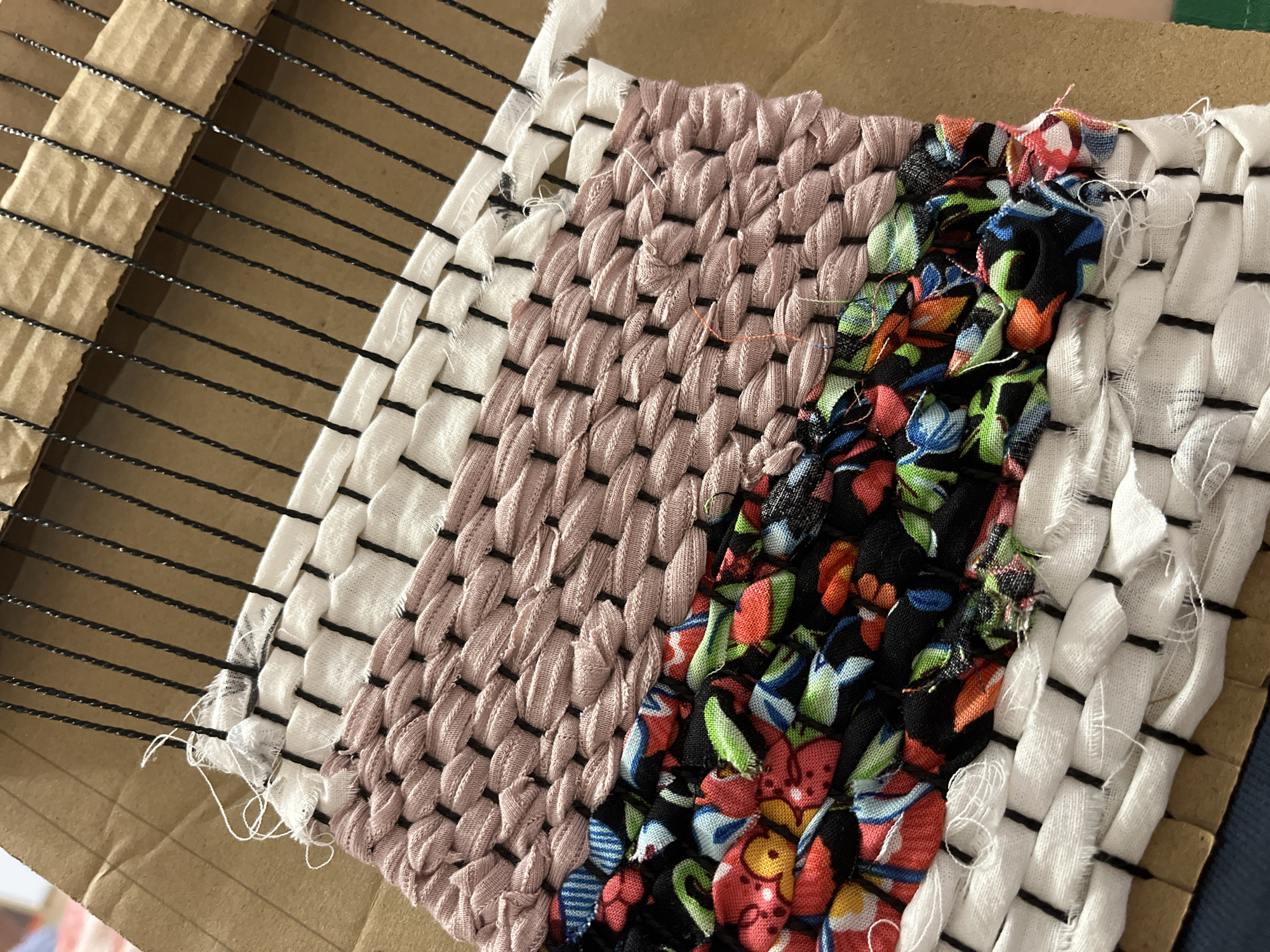 The image shows a close-up view of a small, handmade weaving project on a cardboard loom. The weaving is composed of strips of fabric in various colors and patterns. On the left, white fabric with thin black stripes is woven with a loose, horizontal pattern. Next to it is a section of solid pink fabric, woven in a more intricate, textured pattern. The center of the weaving features brightly colored fabric with a floral pattern, predominantly black, red, blue, and green. To the right of the floral section is another strip of white fabric with black stripes, woven in a similar pattern to the leftmost section. The weaving is still in progress, with loose threads and unfinished edges visible. The background is a textured cardboard surface, likely serving as a makeshift loom, with thin black cords stretched across it to create the warp. The lighting suggests an indoor setting, possibly a craft or home environment. There is no indication of people, a specific time of day, or weather conditions. No text is visible in the image.