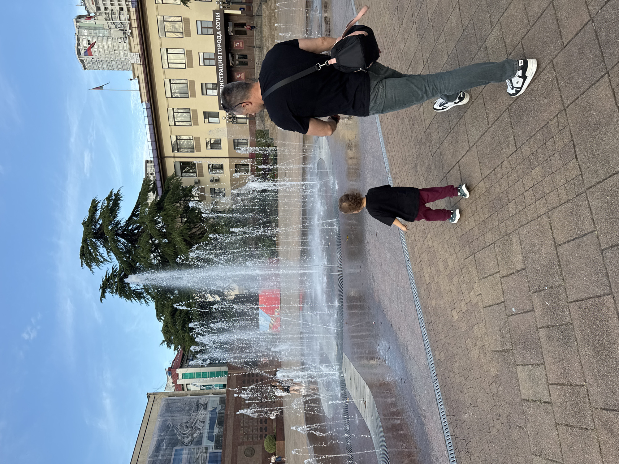 An outdoor scene captures an adult and a child by a modern, ground-level fountain in a city plaza. The image is oriented sideways, with the ground appearing as a vertical plane on the right and the sky on the left.
The adult, viewed from behind, wears a black t-shirt, dark grey jeans, and black-and-white sneakers. A black shoulder bag with a pink strap is visible across their back. They are positioned to the right of the fountain, seemingly observing it or the child. The child, smaller in stature and also viewed from behind, wears a dark top and maroon pants with white sneakers. They stand closer to the fountain's jets, facing towards them.
The fountain features numerous vertical water jets spraying upwards from a paved area. The ground around the fountain is made of brown and grey square tiles, with drainage grates visible along the edge of the fountain.
In the background, under a bright blue sky with a few faint clouds, stand several buildings. Most prominent is a light brown, multi-story building with numerous windows, which features the text "АДМИНИСТРАЦИЯ ГОРОДА СОЧИ" (Administration of the City of Sochi) written in Cyrillic script. Above this building, a tall, modern high-rise is visible, with a Russian flag flying from its roof. Another Russian flag is seen on a flagpole in front of the "Administration" building. A large evergreen tree with dark green foliage is also present on the left side of the frame, partially obscured by the fountain's spray.
The scene depicts a clear, bright daytime, suggesting pleasant weather suitable for outdoor activities.