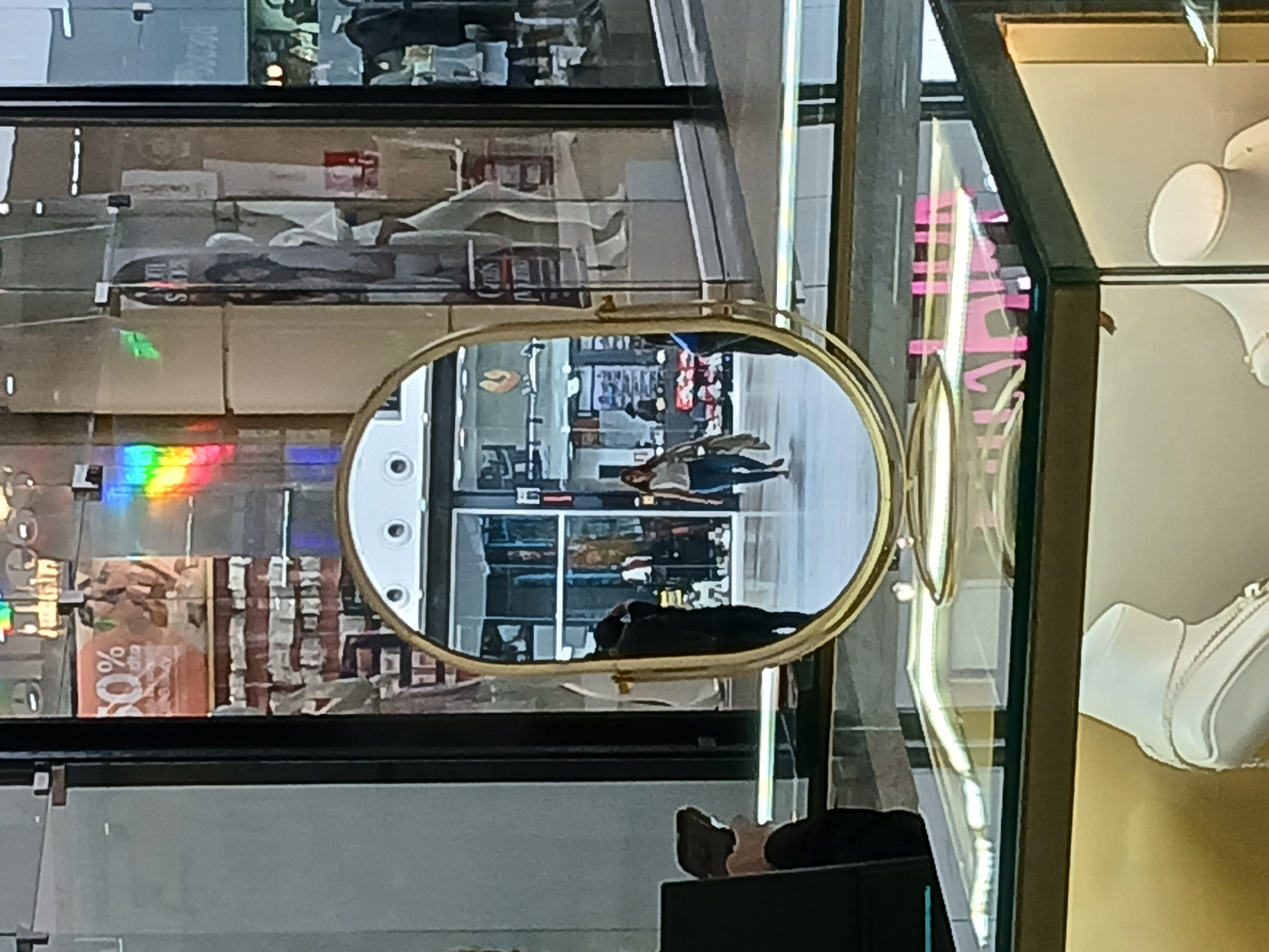 The image shows a shopping mall in Guayana City, Venezuela, reflected in an oval-shaped mirror. The mirror's frame is gold. In the reflection, a couple walks through a mall entrance. The mall is crowded with shoppers and various store displays. To the right of the mirror is a display of white necklaces in a glass case. The image is taken indoors and has a slightly blurred focus. Part of the photographer's body is visible at the bottom. A rainbow reflection appears on the left side of the mirror's surroundings. A "50%" off sign is also visible behind the mirror.