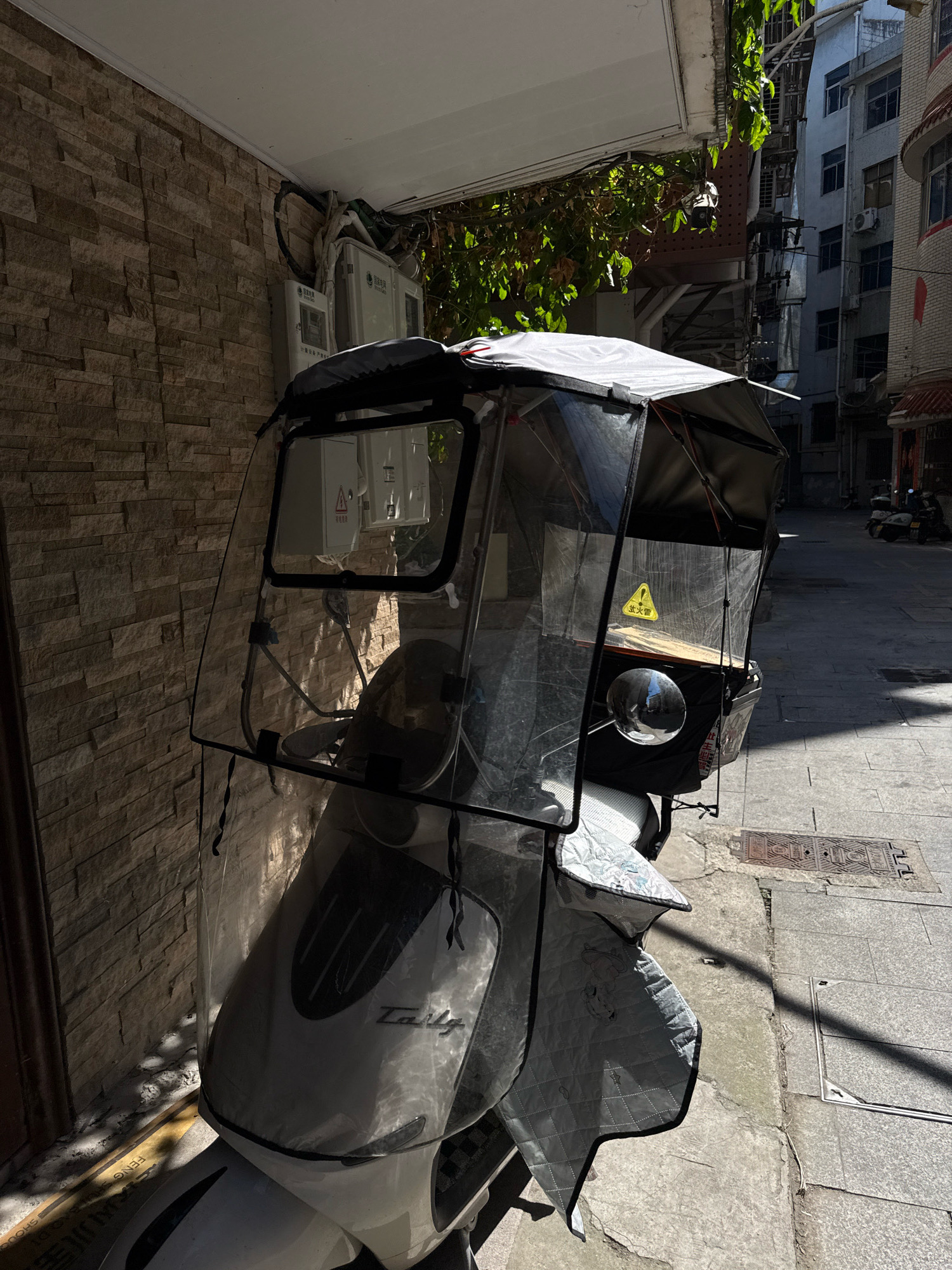A three-wheeled scooter with a clear plastic enclosure and a fabric cover is parked on a paved street in front of a brick wall. The scooter has a white body and a black seat. The enclosure protects the rider from the elements, and the fabric cover is draped over the lower part of the scooter. The street is lined with buildings, and there are some trees and electrical boxes visible. Sunlight is casting shadows on the ground and the scooter. A yellow triangular warning sign with Chinese characters is attached to the inside of the enclosure. There are no people visible in the image.