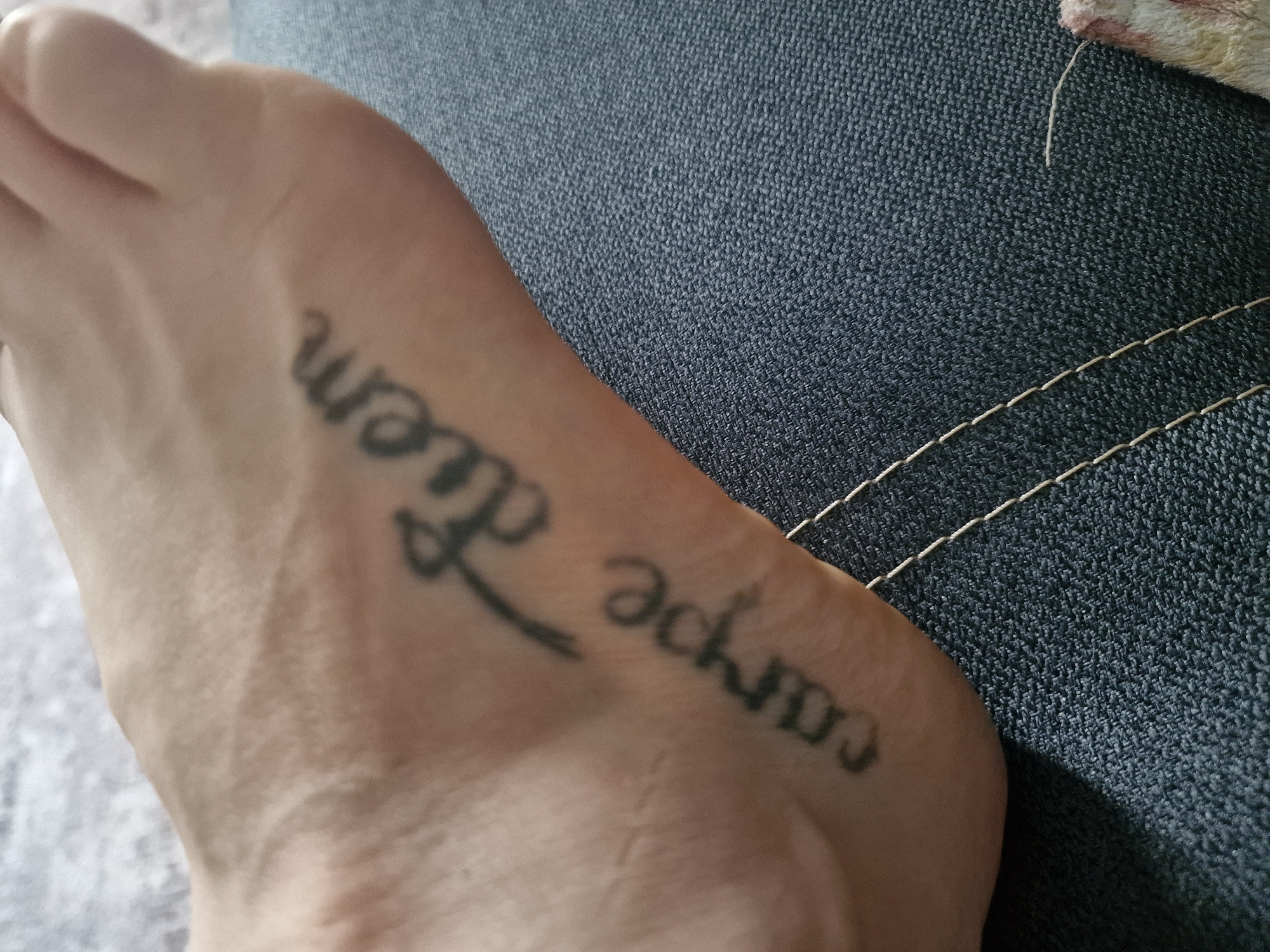 The image shows a close-up view of a person's foot with a tattoo on the top of it. The tattoo is in black ink and appears to say "carpe diem" in a cursive font. The foot is resting on a dark blue denim fabric. There are no discernible city landmarks or environmental features visible that would indicate the location in Kursavka, Russia. The image is tightly cropped and focuses solely on the foot and the tattoo.