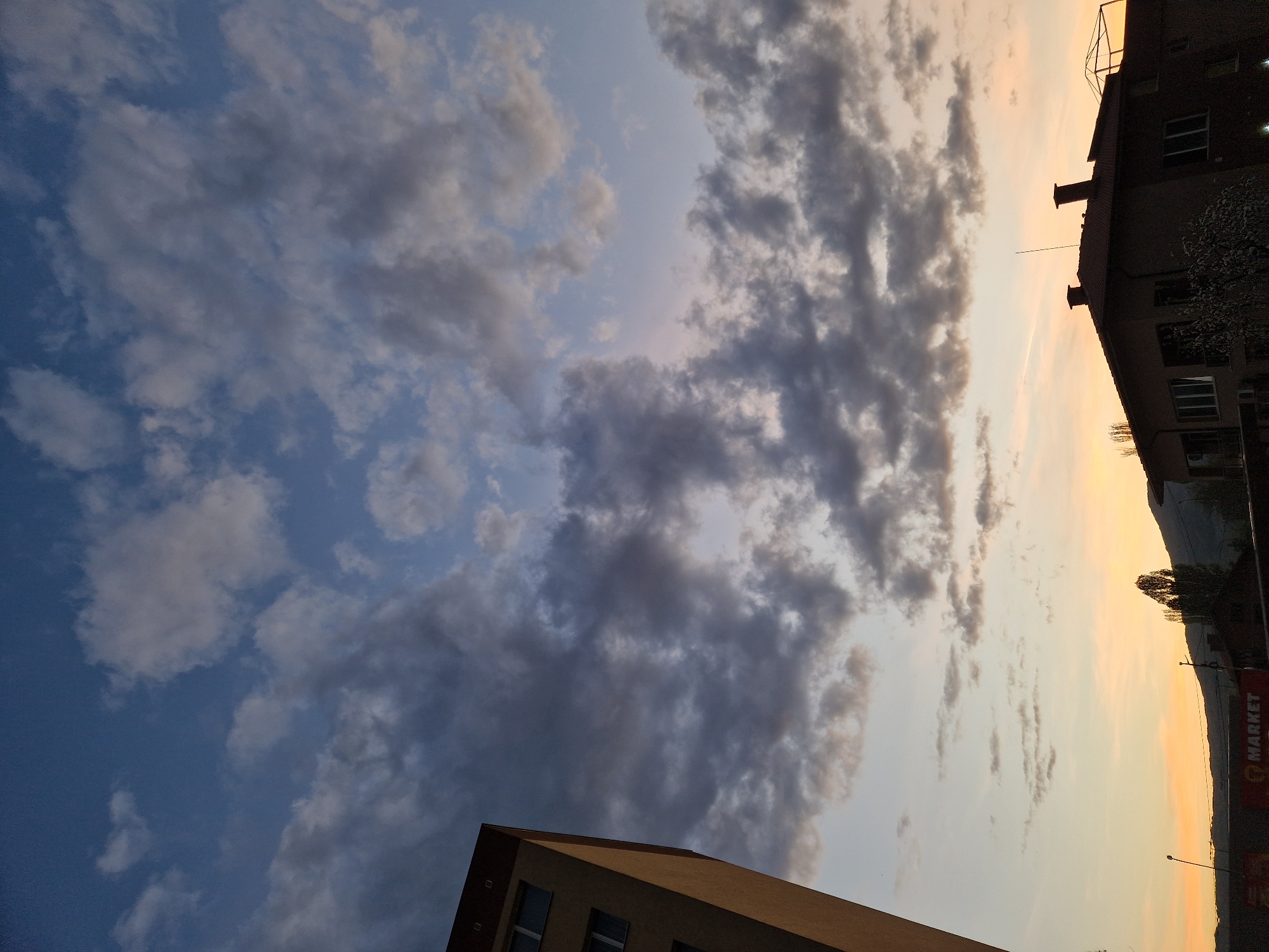 The image shows a view of the sky with clouds in the city of Unknown, Unknown. Buildings are visible on the bottom right and bottom left of the frame. The sky ranges from a light blue to a pale orange-yellow. A sign is partially visible on the right of the frame reading "MARKET". There are trees silhouetted against the sky in the distance.