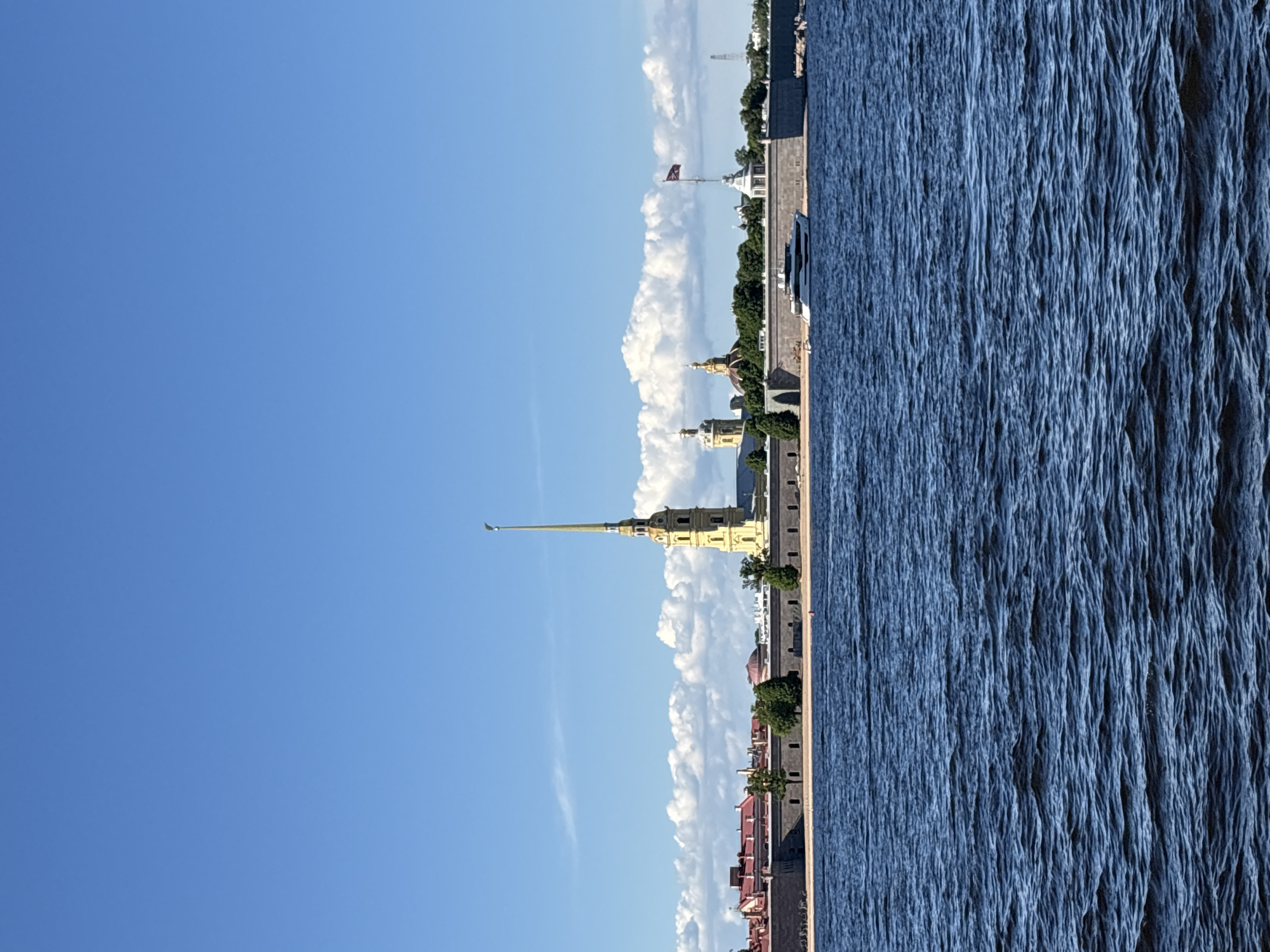 This image depicts the Peter and Paul Fortress in St. Petersburg, Russia, viewed from across the Neva River under a clear blue sky with scattered white cumulus clouds. The lower half of the frame is dominated by the dark blue, rippled surface of the river. The fortress occupies the middle ground, showcasing its grey stone walls, a narrow sandy bank, and interspersed green trees. The prominent golden spire of the Peter and Paul Cathedral rises centrally above the fortress walls, flanked by red-roofed buildings to the left and other golden-domed structures to the right. A dark flag is visible on a flagpole atop the fortress wall to the right. A white luxury yacht is moored alongside the fortress wall, slightly right of center. No individuals are visible.