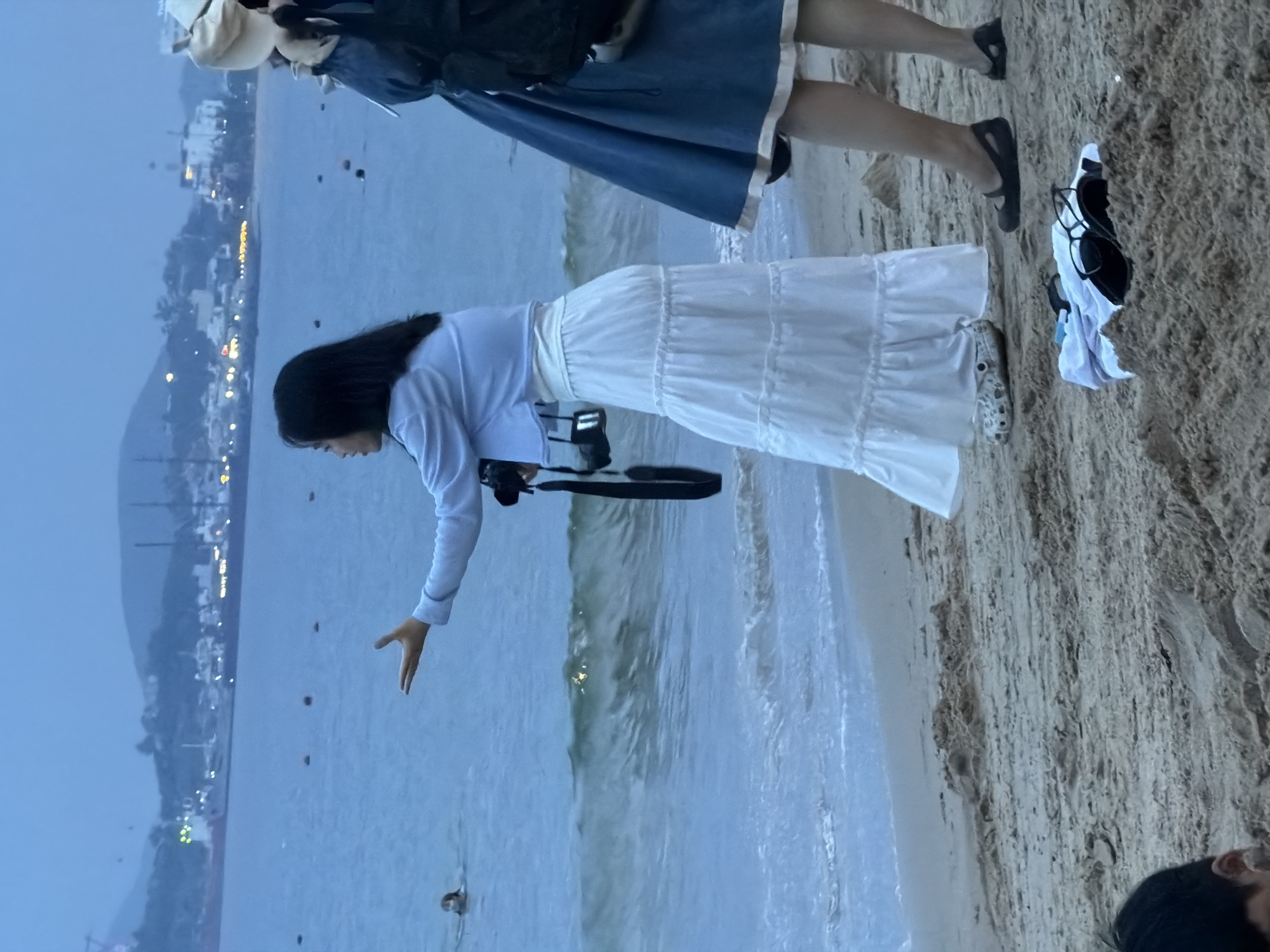 The image depicts a beach scene at dusk in Nha Trang, Vietnam. The foreground is sandy, with a pile of clothing and sunglasses in the center. Two women are visible on the beach. The woman on the left, wearing a light blue jacket and a white tiered skirt, holds a camera and gestures with her left hand towards the sea. She is also wearing patterned clogs. The woman on the right, wearing a blue dress and a hat, stands with her legs crossed. In the middle ground, waves crash onto the shore. In the background, the sea stretches to the horizon, with a coastline featuring buildings and hills dimly illuminated by lights. A person is visible swimming in the water. Several buoys are also present in the sea. The sky is overcast and a muted blue.