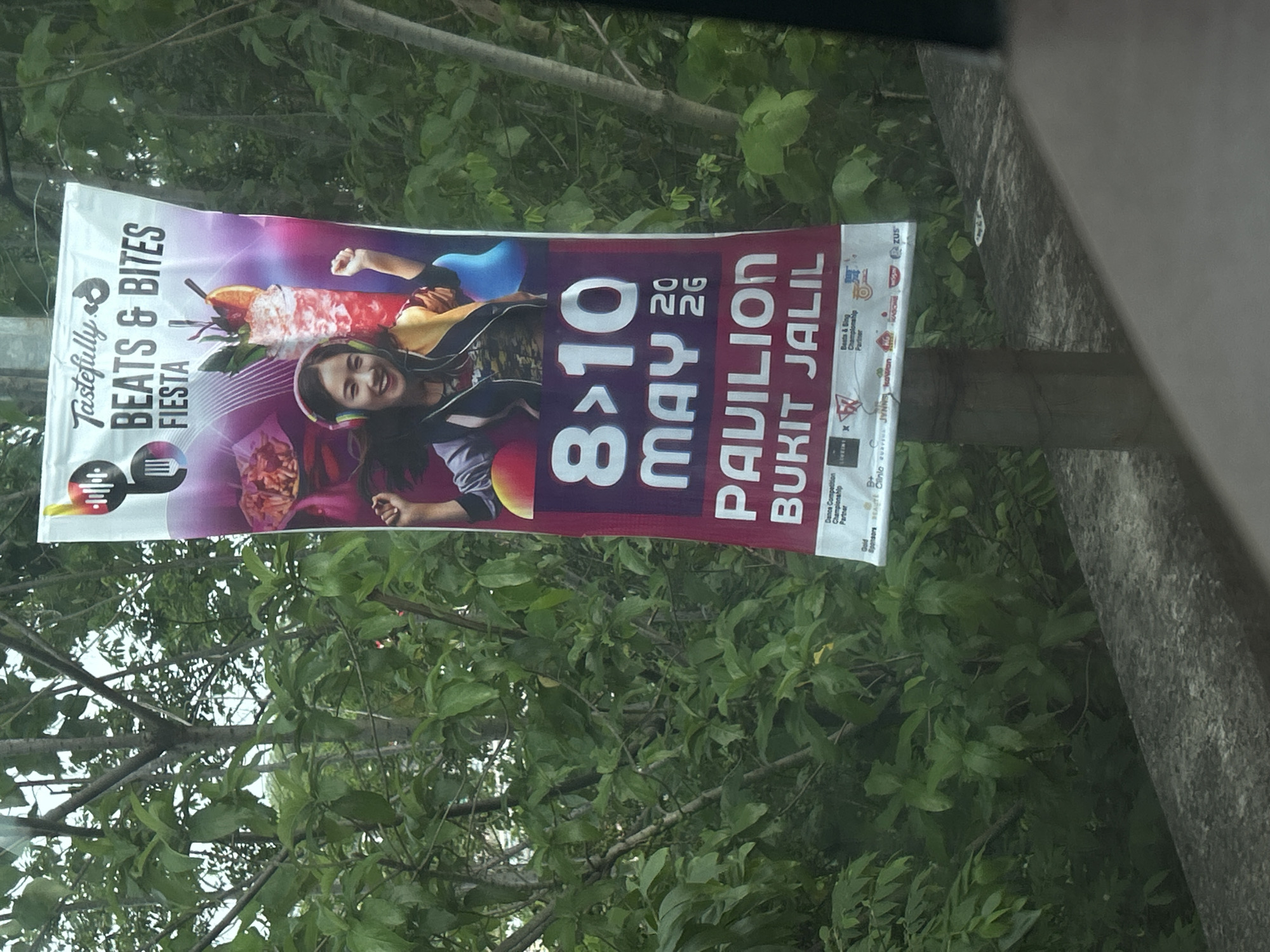 A banner advertises the "Tastefully Beats & Bites Fiesta" event, taking place from May 8 to 10, 2020, at Pavilion Bukit Jalil in Kuala Lumpur, Malaysia. The banner features a young woman with headphones on, seemingly enjoying music. Sponsors for the event are listed at the bottom of the banner. The banner is affixed to a post against a backdrop of lush green foliage.