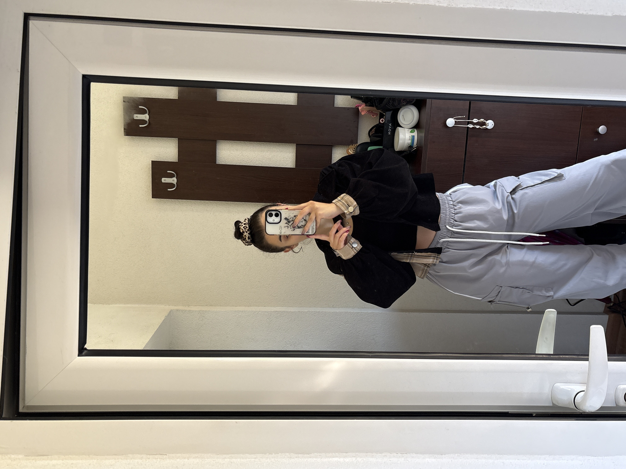 A young woman takes a selfie in a mirror. The reflection shows a room with a coat rack mounted on the wall, a set of dark brown drawers, and toiletries. The woman is wearing a black top, light gray pants, and her hair is in a bun. The location is Polena, Bulgaria, as indicated by the geographical references in the image metadata.