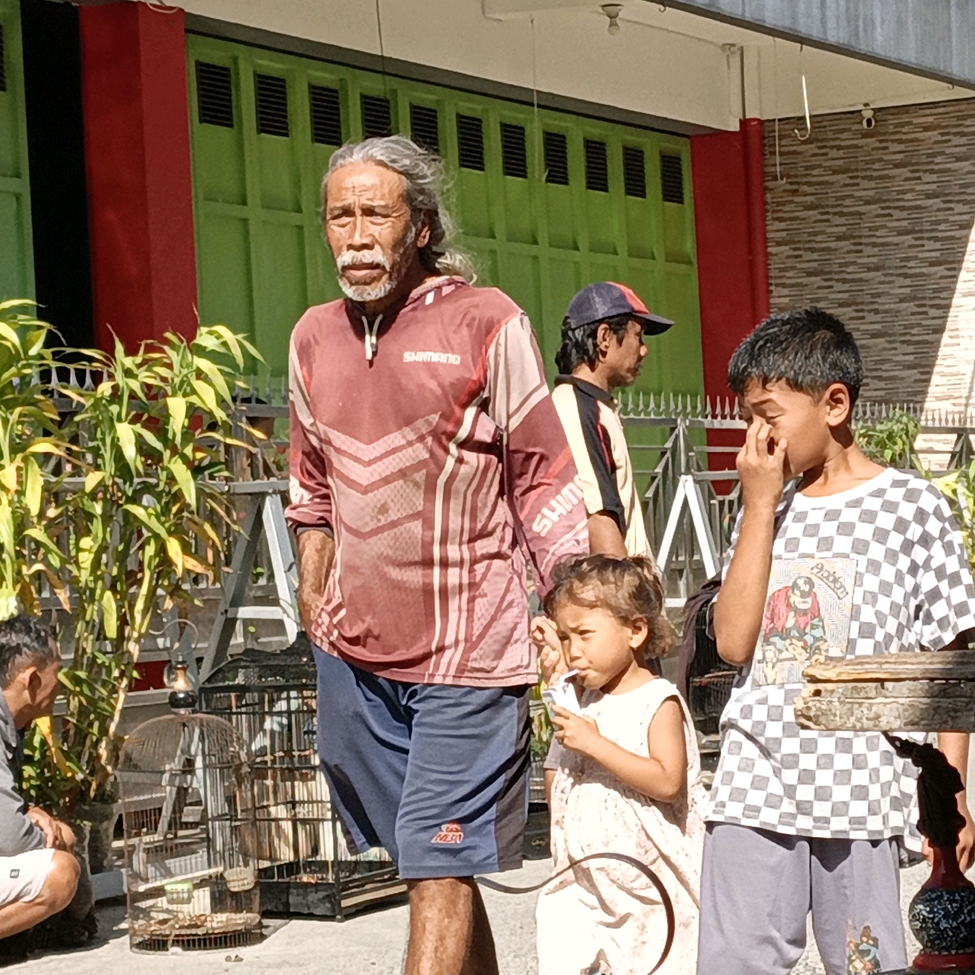 In Randudongkal, Indonesia, an elderly man with graying hair and a beard stands in front of a building with green doors and red columns. He wears a maroon long-sleeved shirt with a geometric pattern and blue shorts. Beside him, a young boy with dark hair wears a checkered t-shirt and gray shorts, and he is touching his face. A young girl in a light-colored dress stands between them, holding a small white object. Behind them, another man wearing a cap is visible. Various bird cages are placed on the ground in front of the building. The scene appears to be outdoors on a sunny day.