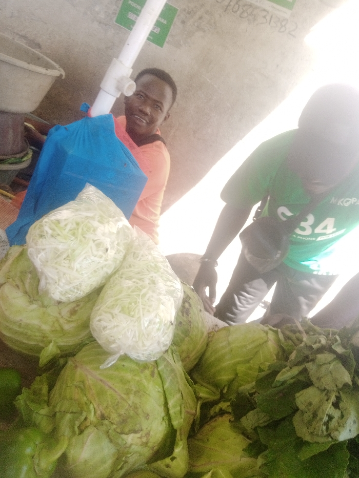 The image depicts a produce stall within a market environment in Mtopanga ward, Kenya. In the foreground, a large quantity of green cabbages is displayed, some appearing whole, while two clear plastic bags containing shredded white cabbage are placed on top of them. To the right, a pile of dark green leafy vegetables is visible, alongside a single light green bell pepper in the bottom left.
Two individuals are present. On the left, a light-skinned male wearing an orange long-sleeved shirt smiles directly at the camera. He is positioned behind a blue fabric bag and some produce. To his right, partially obscured from the torso down, a dark-skinned male wears a green t-shirt with "MKOPA" and "C34 A PHONE" printed on it, along with dark trousers. He is leaning forward with his right hand near the produce.
In the background, a white PVC pipe structure extends vertically. Behind the smiling male, a large white plastic basin is visible. A light-colored wall or ceiling surface shows a green sign with partial text "POCHA" and a phone number "768431382". The lighting is bright and diffuse, consistent with an indoor or covered market setting.