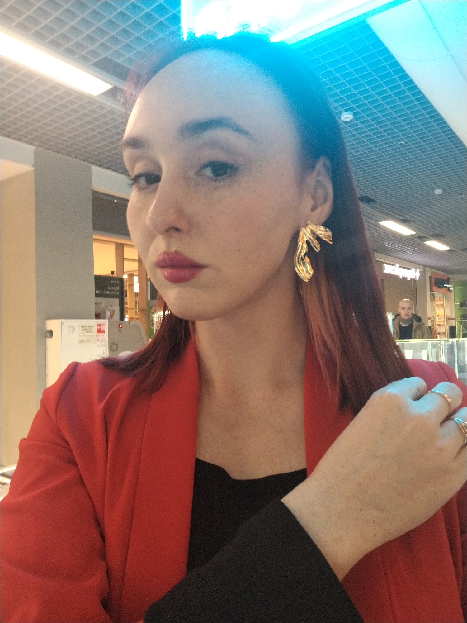 A woman with auburn hair and freckled skin takes a selfie indoors. She wears a red blazer over a black top and has gold statement earrings. She is wearing multiple gold rings on her fingers. Behind her is a man standing in a store. The ceiling appears to be a grid pattern with fluorescent lighting. The photo was taken in Izhevsk, Russia.
