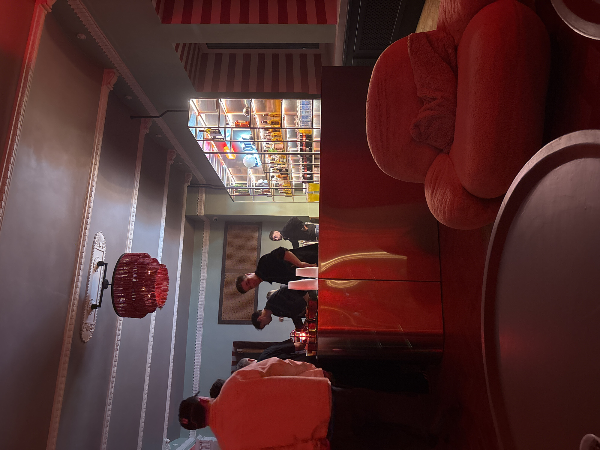 The image depicts the interior of a restaurant or bar. Several people are standing around a bar area, including what appear to be bartenders and patrons. A large red, puffy chair is in the foreground. In the background, there is a well-stocked bar with numerous bottles on shelves. The decor includes striped patterns on the ceiling and decorative molding on the walls. A unique red lamp fixture is visible on the left wall. The location appears to be an upscale establishment.