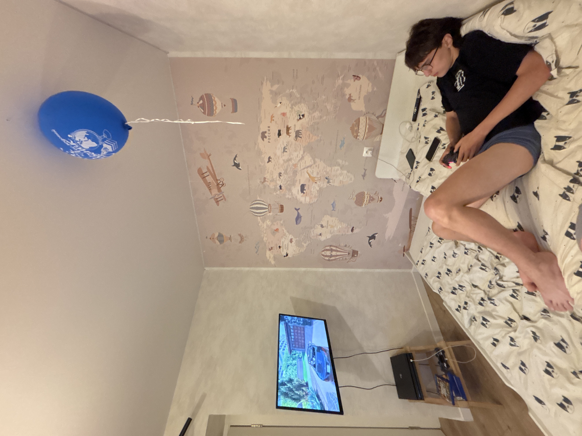 A young person is seen relaxing on a bed in an indoor setting, likely a bedroom in Chaikovsky, Russia. The image captures the room from a rotated perspective, with one wall featuring a decorative world map. The individual, appearing to be a young adult or teenager, wears glasses, a dark t-shirt with a white graphic design, and dark shorts. They are lying on their side, holding a dark video game controller, seemingly playing a game or watching content on a flat-screen TV mounted on the opposite wall. A smartphone and a remote control are visible on the patterned duvet, which features small black and white dog faces.
The wall behind the bed is adorned with a large, light-toned world map decorated with various cartoon animals (such as bears, whales, giraffes, and kangaroos) and illustrations of hot air balloons and early airplanes. Russian text on the map clearly shows "РОССИЯ" (Russia) and "АВСТРАЛИЯ" (Australia). Near the ceiling, a blue balloon with a white string floats, bearing white text that partially reads "Graduation" and an emblem of a globe with a cap, suggesting a celebratory occasion. A small wooden shelf next to the bed holds a black laptop or similar electronic device, with visible cables connecting to the television. The lighting in the room is soft and ambient, indicative of an indoor environment without strong natural light. The scene depicts a moment of casual leisure and entertainment.