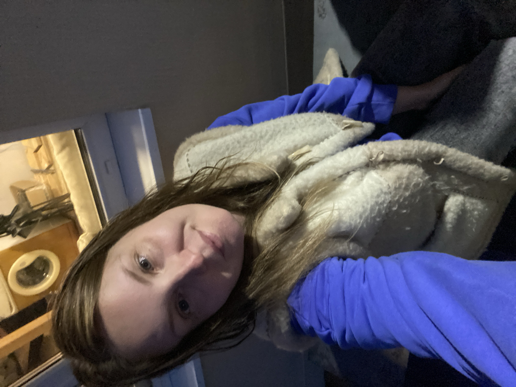 A woman with long, light brown hair is sitting in a room, possibly an attic, given the sloped ceiling and window. She is wearing a bright blue hooded sweatshirt and a fuzzy cream-colored vest with toggle closures. Her hair is partially covering her face and shoulders. The background shows a window with a view of what appears to be a wooden washing machine or dryer in a dimly lit space with exposed wooden beams. The floor is covered with a dark fabric or carpet. The overall lighting suggests it is evening or indoors. The location is identified as Garevaya, Russia.