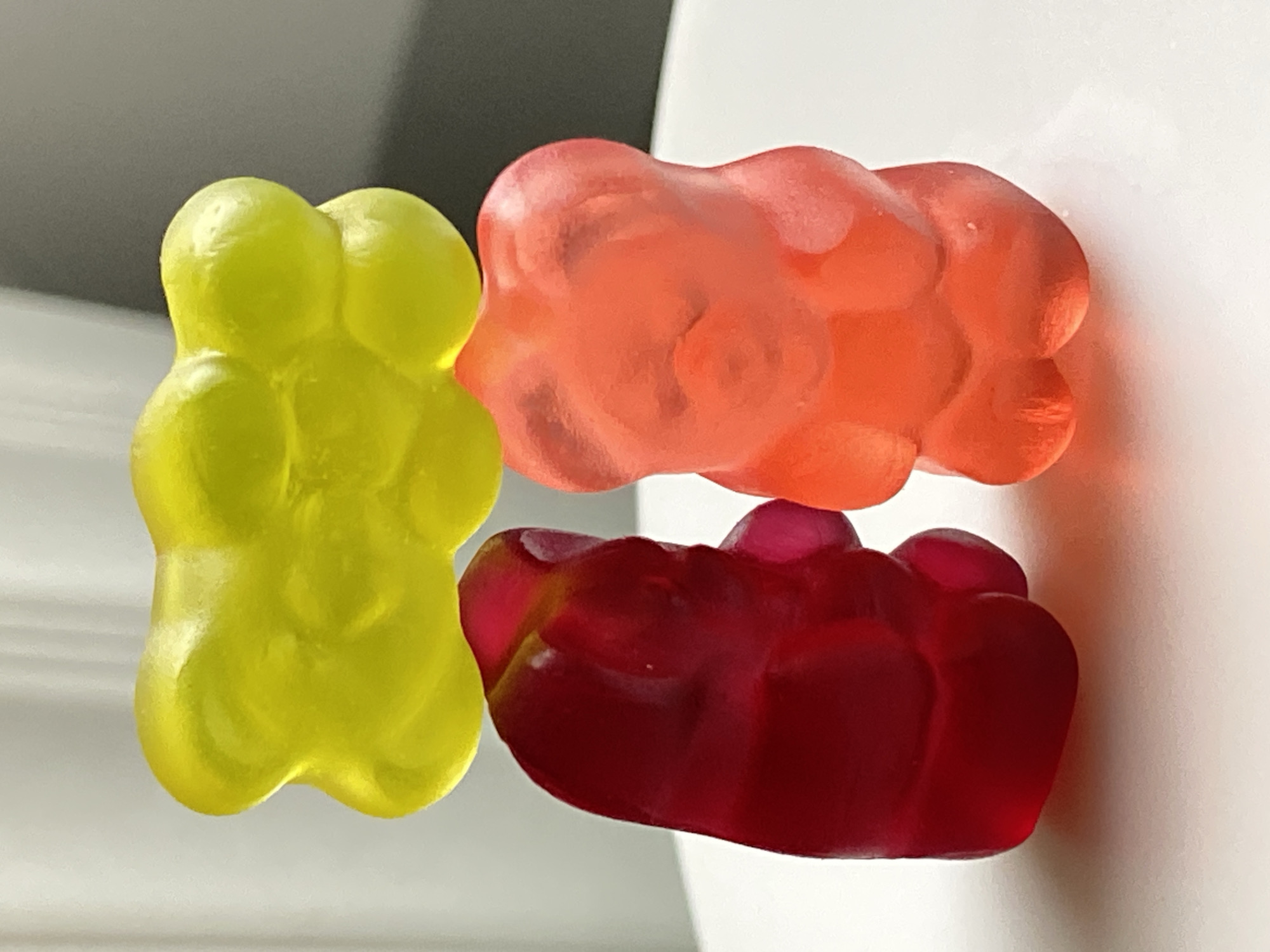 A close-up photograph displays three translucent gummy candies against a soft, blurred background. On the left, a bright lime-green or yellow gummy stands upright, clearly identifiable as a gummy bear with distinct features. To its right, an orange-red gummy is positioned horizontally, appearing slightly squashed or melted, resting partially on a deeper red or maroon gummy below it. This third gummy also lies horizontally and has an indistinct, rounded shape.
All three candies exhibit a glossy, somewhat sticky texture with highlights reflecting the ambient light. The background is predominantly light-colored and out of focus, with a curved white object visible on the right, possibly a cup or mug. Some darker, blurred areas in the upper left suggest depth and contribute to the abstract setting. The lighting is even and bright, emphasizing the vibrant colors and soft, pliable nature of the confections. No activity is occurring, and there is no visible text.