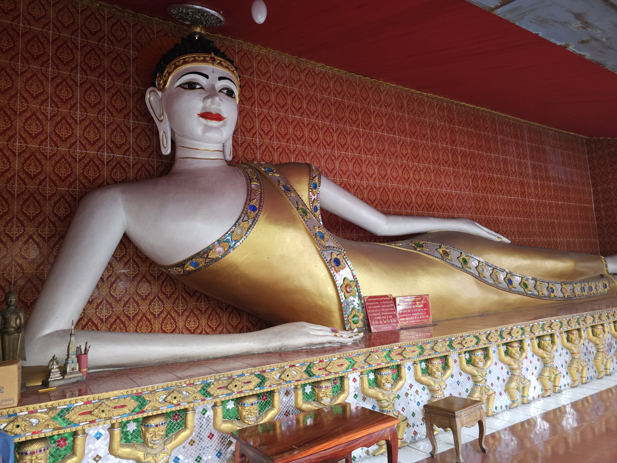 The image shows a large, reclining Buddha statue in a temple. The statue is white with gold robes and is adorned with colorful jewels. The background is a red, tiled wall. In the foreground, there is a wooden table and a small stool. The floor is tiled in a checkerboard pattern. The statue is lying on a raised platform decorated with golden figures. The overall impression is one of grandeur and serenity. This scene is typical of many Buddhist temples in Bangkok, Thailand, where such statues are common.