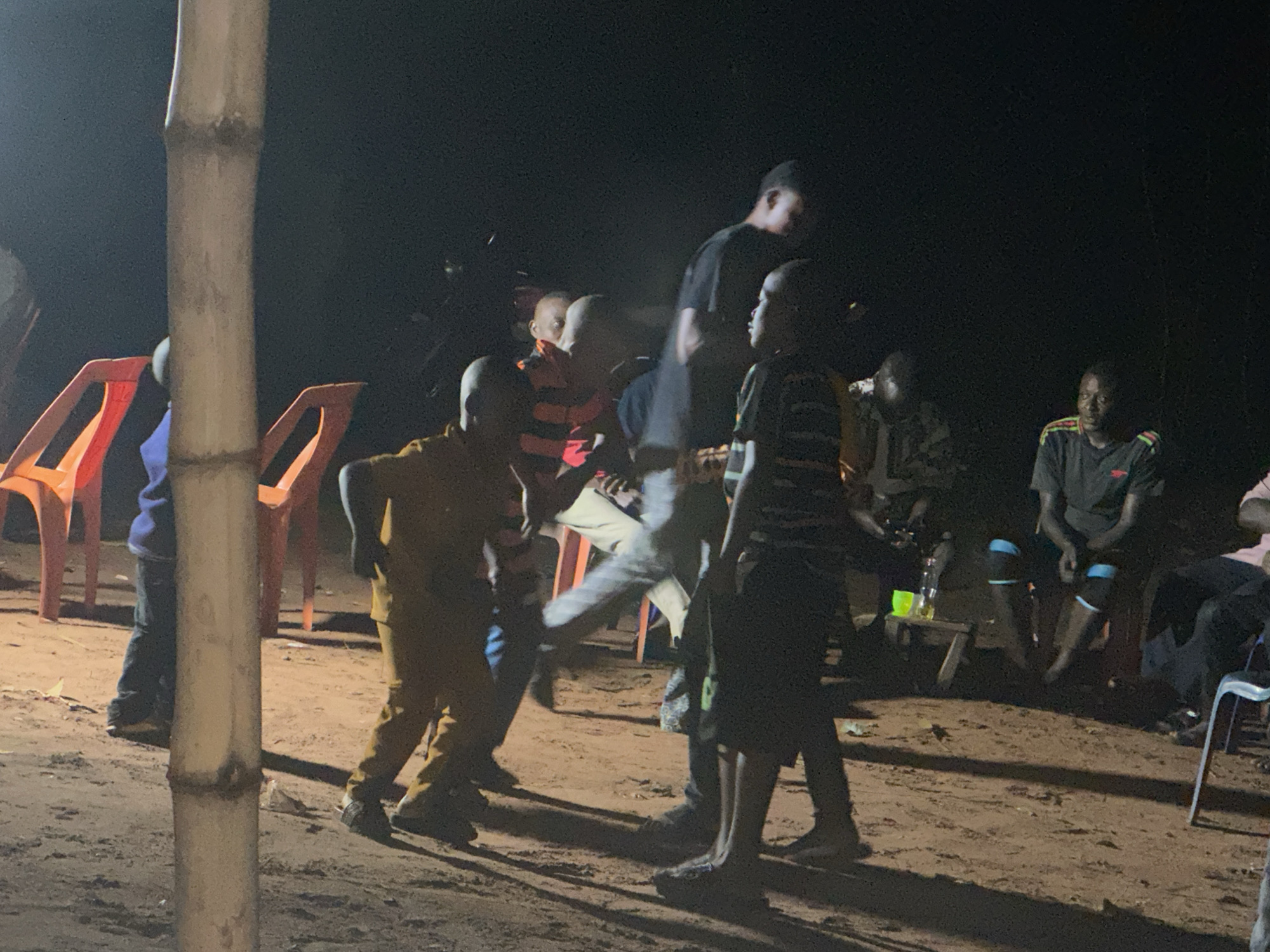 The image shows a group of people gathered outdoors at night. The ground appears to be dirt or sand. In the foreground, there is a pole made of light-colored segments. Several orange plastic chairs are visible. Some people are standing and appear to be moving, while others are sitting on chairs or benches. A small wooden table with bottles and cups on it is present. The scene is illuminated by artificial light, but the source is not visible. The location is Unknown, Unknown.