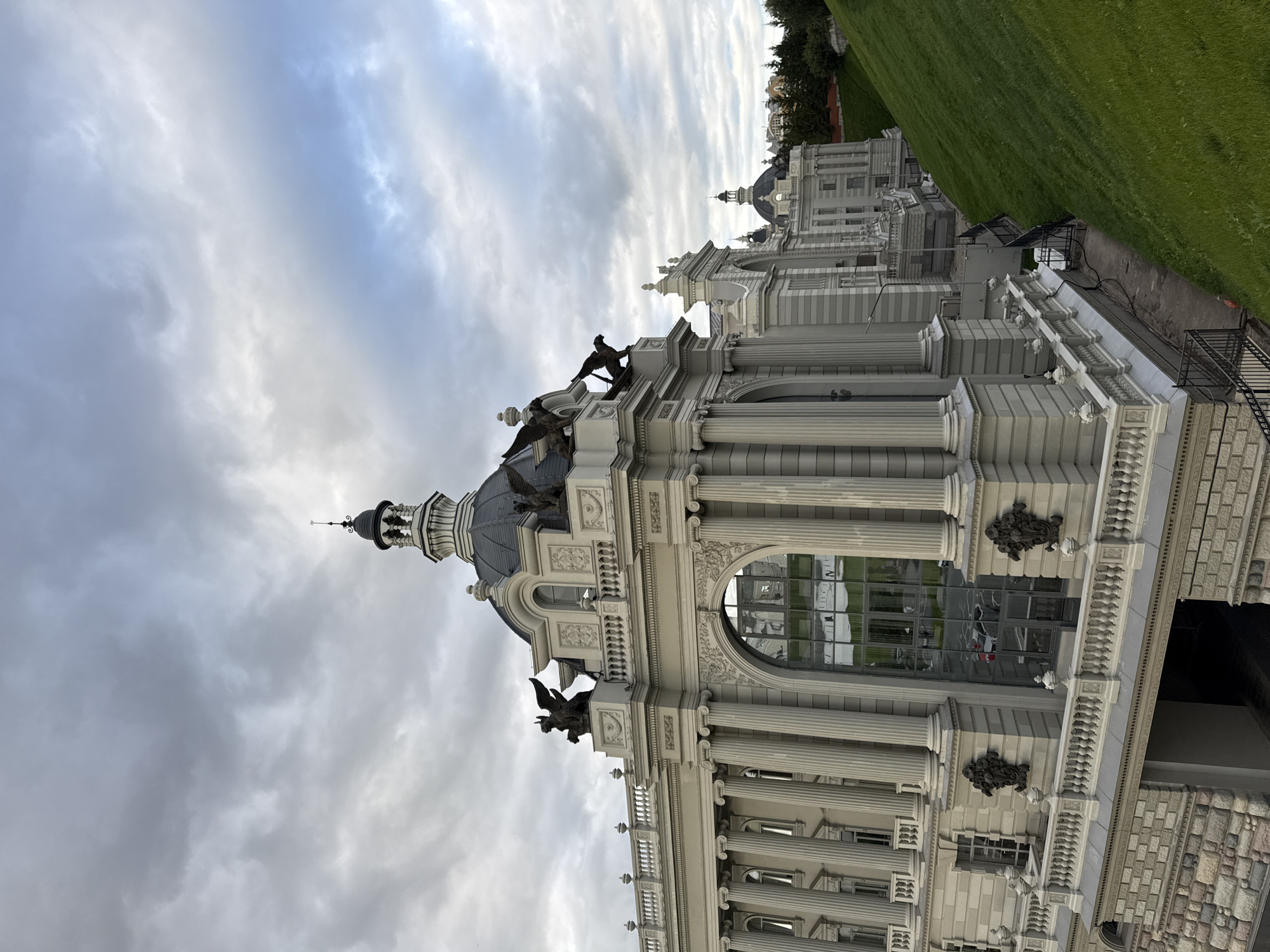 The image depicts the main facade of the Palace of Agriculture, a prominent architectural landmark in Kazan, Russia. The building is characterized by its neoclassical design, featuring large Corinthian columns, a grand arched entrance with a glass facade, and decorative sculptures of griffins atop the roofline. The building is adorned with ornate details, including balustrades and decorative motifs. A steep, green grassy embankment slopes down to the right of the building, creating a strong contrast between the man-made structure and the natural landscape. The sky above is overcast with dramatic grey clouds, suggesting a temperate climate. The overall scene is one of grandeur and historical significance, highlighting the architectural splendor of the Palace of Agriculture.