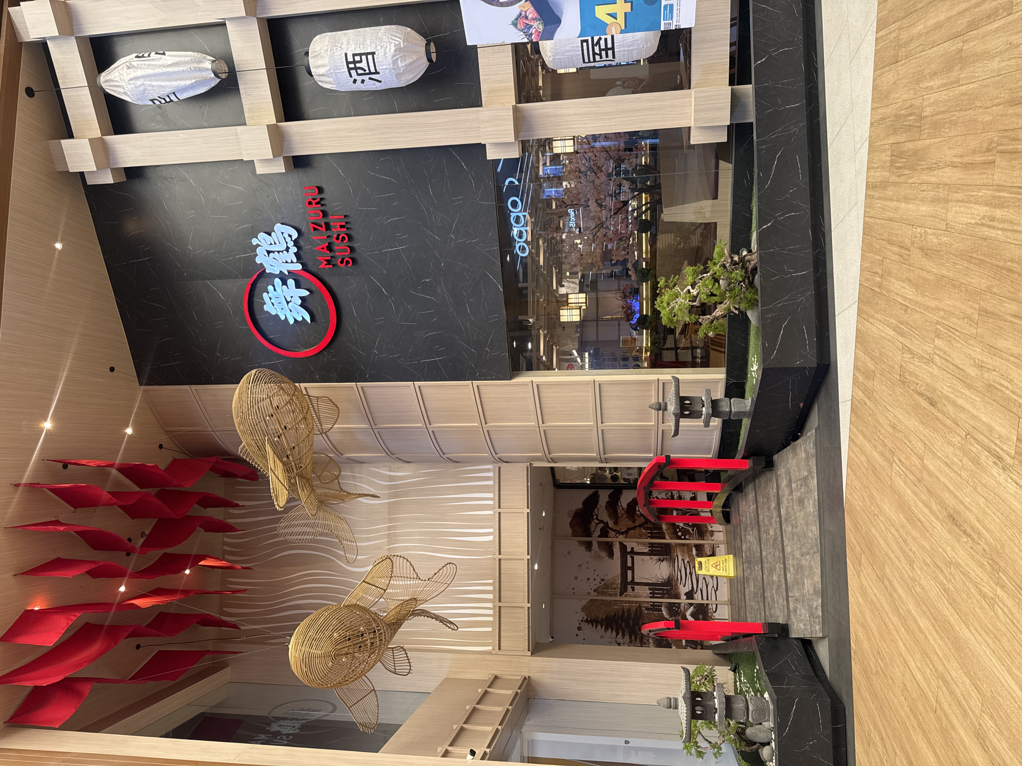 The image shows the entrance of a Japanese restaurant named "Maizuru Sushi" in a mall. The restaurant's facade features a black marble wall with the restaurant's logo in red and white. Above the logo, there are three Japanese lanterns hanging. The restaurant's entrance has a small Japanese garden with a stone lantern and a red torii gate. The interior of the restaurant is visible through the glass doors. There are two large golden fish sculptures hanging from the ceiling, as well as red decorations that resemble fish scales. The location appears to be in a modern shopping mall, as evidenced by the flooring and architectural details. Based on the location, it is likely in the city of Kuta, Indonesia.