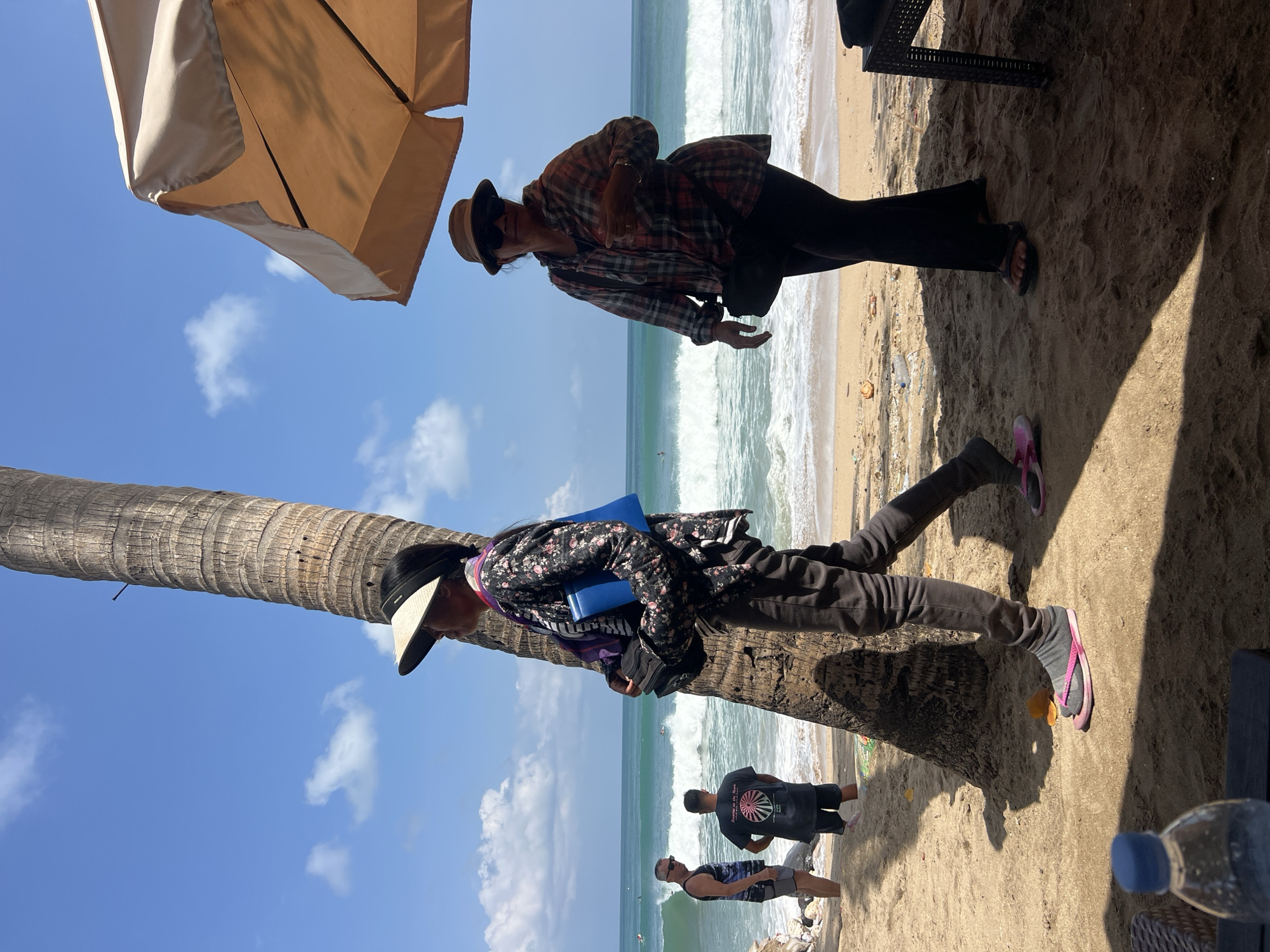 In Kuta, Indonesia, on a sunny day at the beach, two women are seen near a palm tree and a large umbrella. The woman on the left, partially obscured by the palm tree, wears a floral print jacket, dark pants, and pink flip-flops. She is carrying a blue bag and what appears to be a stack of dark items. The woman on the right, standing under the umbrella, wears a plaid shirt, dark pants, and sandals. She is looking towards the camera and has sunglasses on. In the background, the ocean has moderate waves, and several people are visible on the beach and in the water, some appearing to be surfers. The beach is sandy with some debris scattered around.