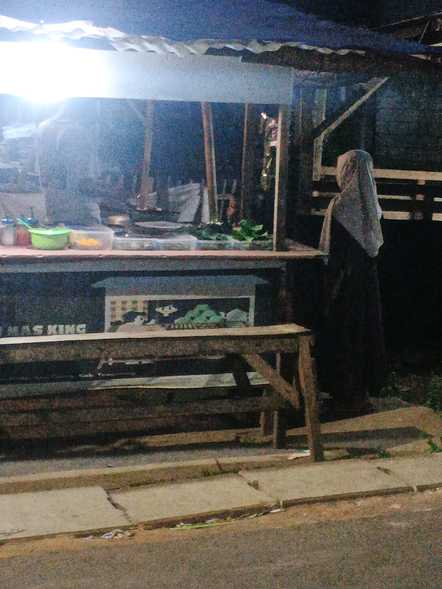 The image depicts a street food stall at night in Kadu, Indonesia. The stall appears to be a temporary structure with a corrugated metal roof supported by wooden posts. A bright light illuminates the front of the stall, casting shadows. On the counter, there are various food items, including containers with what appear to be ingredients or prepared food, and bottles of condiments. A person, partially visible and wearing a headscarf, stands behind the counter, seemingly attending to a customer or preparing food. Another figure is visible in the background, also behind the counter. In front of the stall, there is a wooden bench for seating. The stall is situated on a paved street next to a sidewalk. The overall atmosphere is that of a typical late-night food vending operation in a local Indonesian setting.