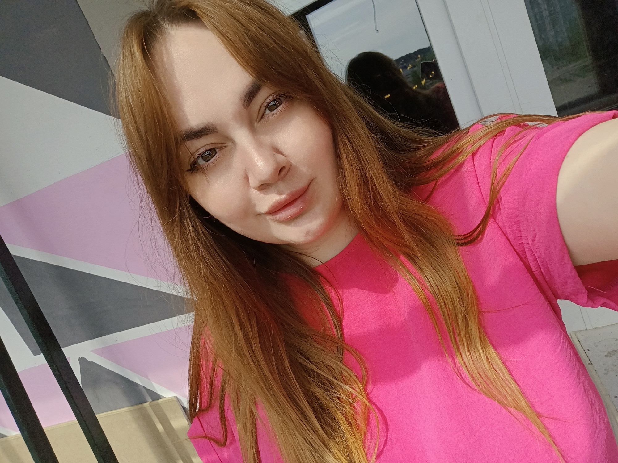 A young woman with long, reddish-brown hair is taking a selfie. She is wearing a bright pink t-shirt. The background includes a wall with a geometric design of grey, white, and pink triangles, and a window reflecting an outdoor scene, possibly of the city of Chaikovsky, Russia, based on the prompt. The reflection shows buildings and some greenery.