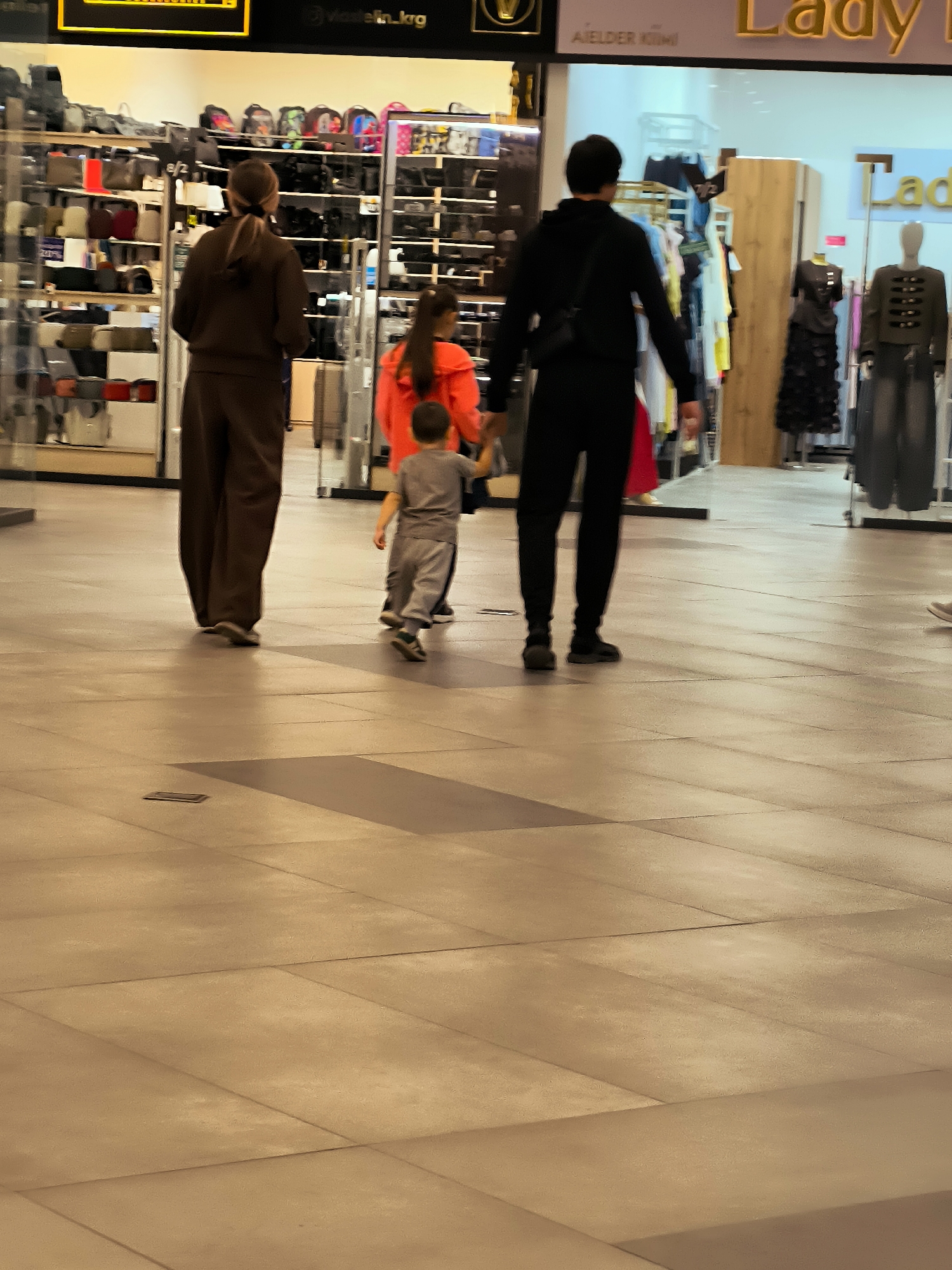 In a shopping mall in Qarağandı city administration, Kazakhstan, a family walks through a retail area. A woman with her hair in a ponytail, wearing a brown tracksuit, walks away from the camera. Behind her, a young girl in an orange top and a young boy in a grey tracksuit are walking, holding hands. Further ahead, a man in a black tracksuit walks away from the camera. The environment includes shelves stocked with bags and clothing, mannequins displaying outfits, and a tiled floor. The lighting suggests an indoor, commercial setting.