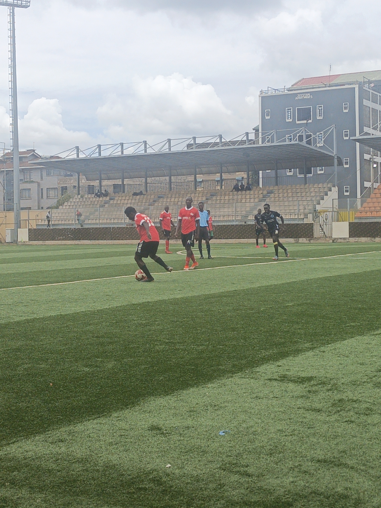 A soccer game is being played on a green field with white lines. Multiple players are visible; some wear red jerseys with "KIPPEN FC" printed on them, while another wears a dark jersey with a white design. One player in a red jersey is dribbling a soccer ball. A referee in a blue shirt stands on the field. Behind the field is a stadium with rows of seats and a metal roof. A tall light pole stands to the left of the stadium. A building with "JOYSTAN APARTMENTS" written on it is visible in the background. The sky is overcast with heavy clouds. The scene appears to be in Nairobi, Kenya.