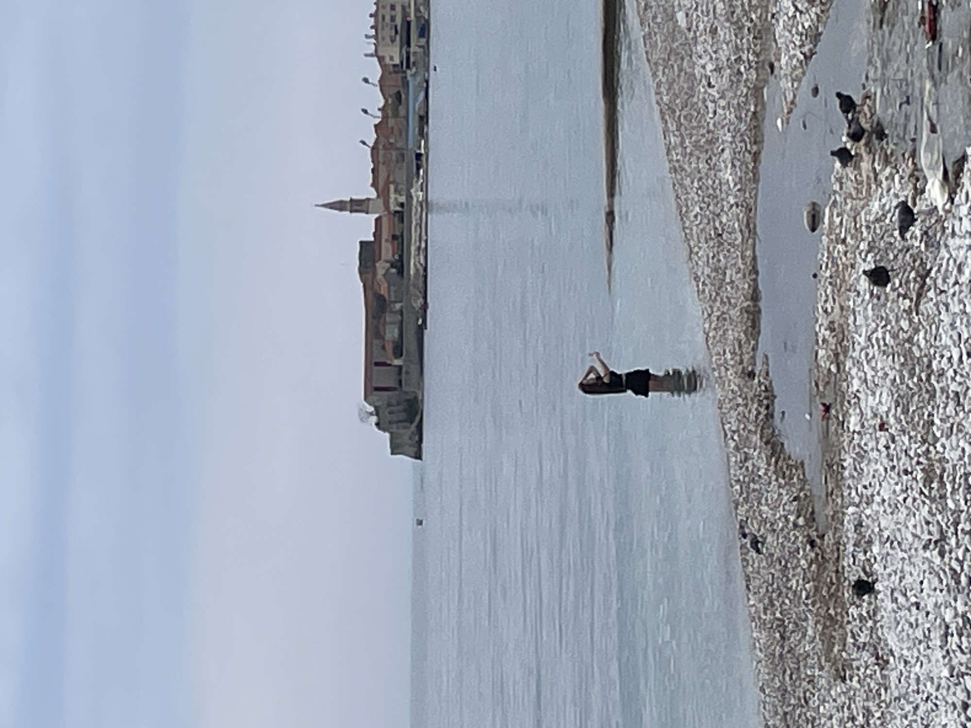 A wide shot captures a cityscape across a body of water, with a swimmer in the foreground.
**Cityscape:** On the left, a cityscape displays a cluster of buildings, including a prominent church spire, typical of coastal settlements. The buildings are various sizes and colors.
**Body of Water:** The scene is dominated by a large body of water with a gray-blue hue, reflecting the sky. The water appears calm, with minor ripples and reflections.
**Swimmer:** A person is swimming in the water, relatively close to the shore. The swimmer is dark, possibly clothed.
**Shoreline:** The shoreline appears to be pebble beach with pebbles of varying sizes and colors. Some dark objects, likely birds or rocks, are scattered along the shoreline.
**Location:** Considering the architecture of the city, the scene likely depicts the coastal town of Budva, Montenegro.