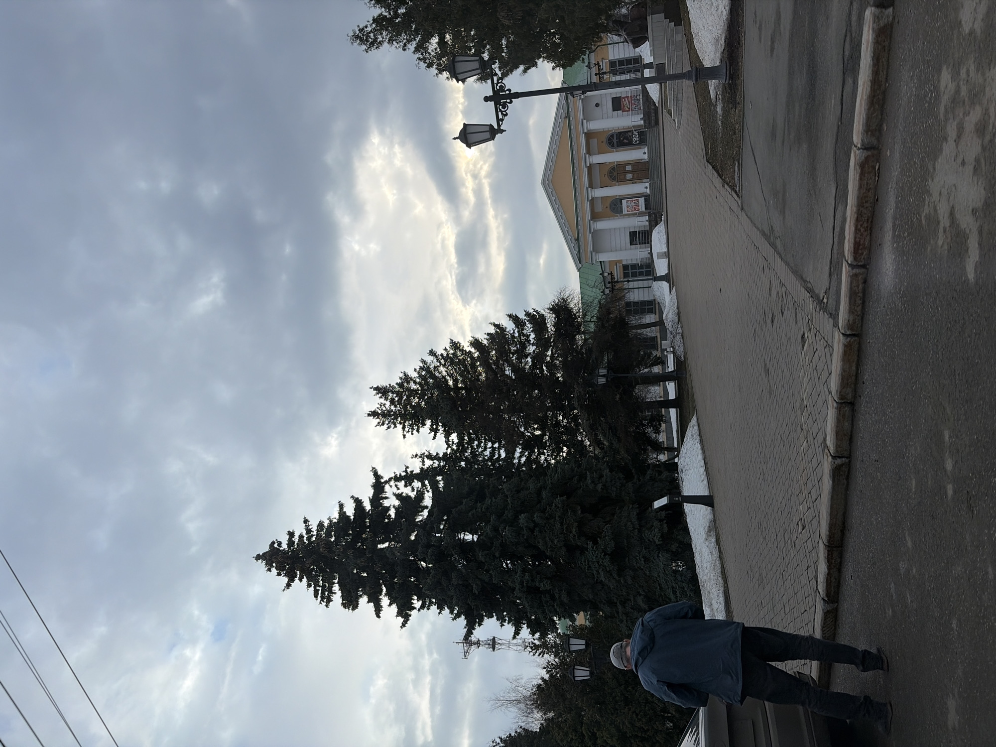The image depicts a street scene in Izhevsk, Russia. A man in a blue jacket and dark pants walks along a paved sidewalk next to a road. Large evergreen trees stand adjacent to the sidewalk. A building with classical architecture, featuring columns and a yellow facade, is visible in the background. Street lamps are placed along the sidewalk. The sky is overcast.