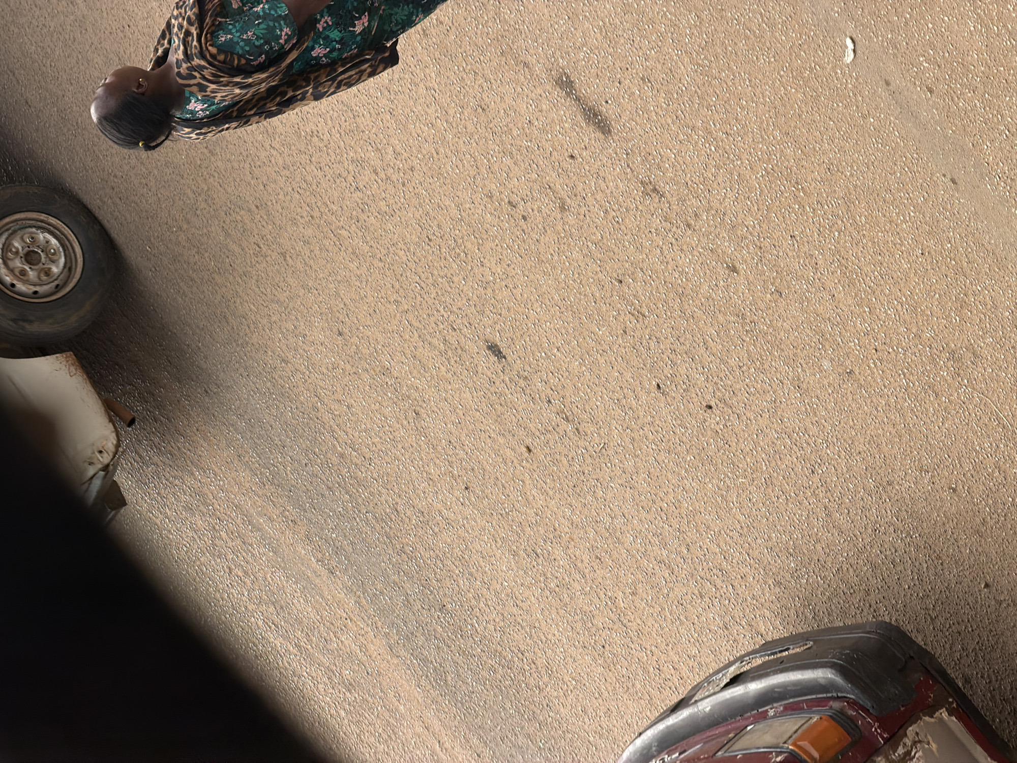 A woman stands on a dusty road in Bukuru, Nigeria, looking upwards. She is wearing a floral green dress with a leopard print scarf draped over her shoulders. Her hair is pulled back in a bun, and she has a small earring in her left ear. Beside her, a portion of a car is visible. The car is dark in color, with a visible headlight and bumper. In the background, another car is partially visible, with a tire and part of its body. The ground is covered in sand and small stones, suggesting an arid or dusty environment. The scene appears to be captured during daylight, with shadows visible on the road.